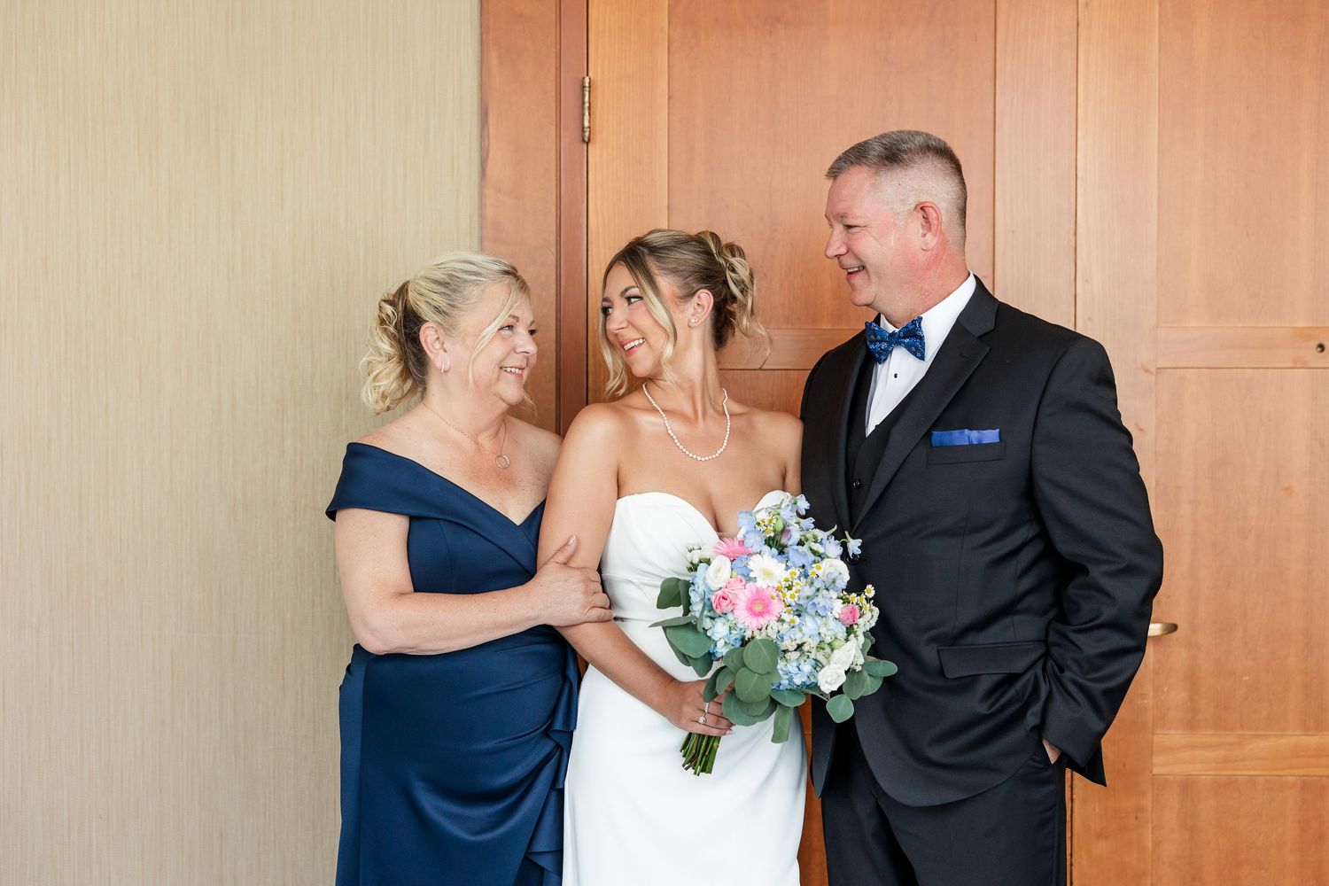 Bride holding floral bouquet posing with parents in navy dress and black tuxedo before wedding.