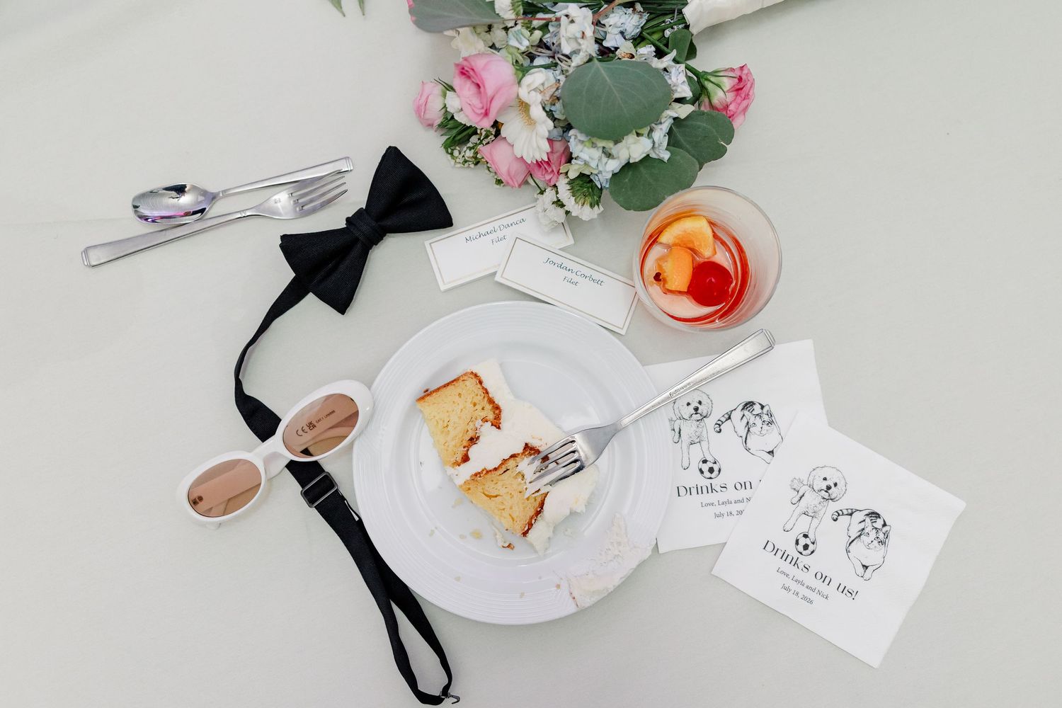 Flat lay of wedding cake slice, black bow tie, sunglasses, florals, and custom favor cards.