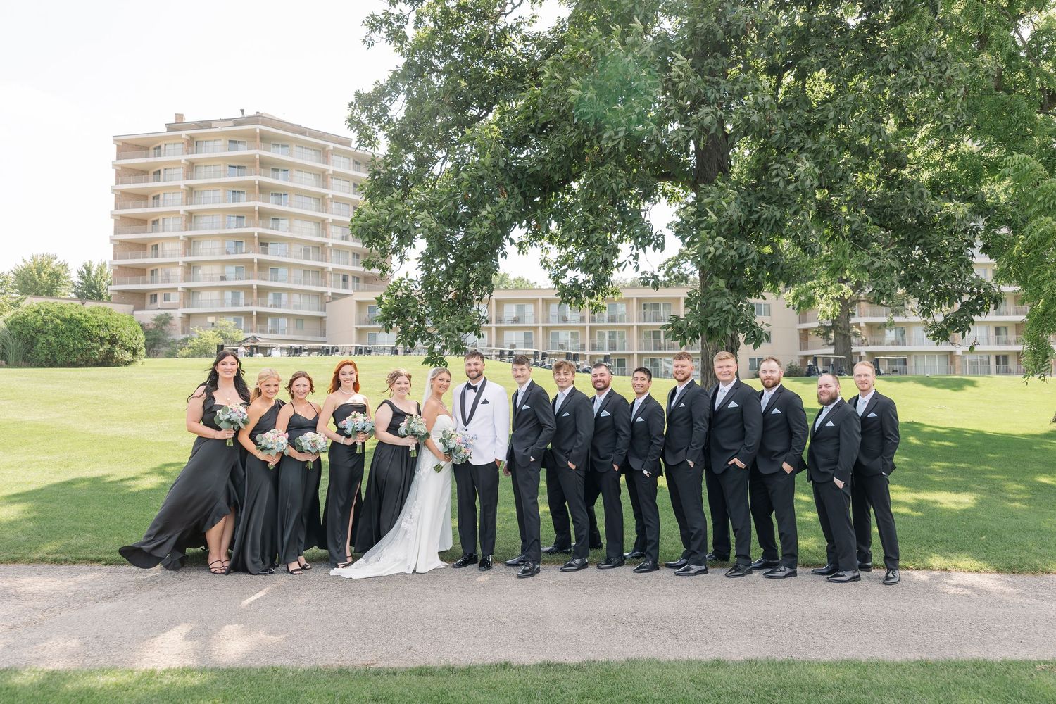 Large wedding party in black attire posing outdoors on green lawn with hotel in background.