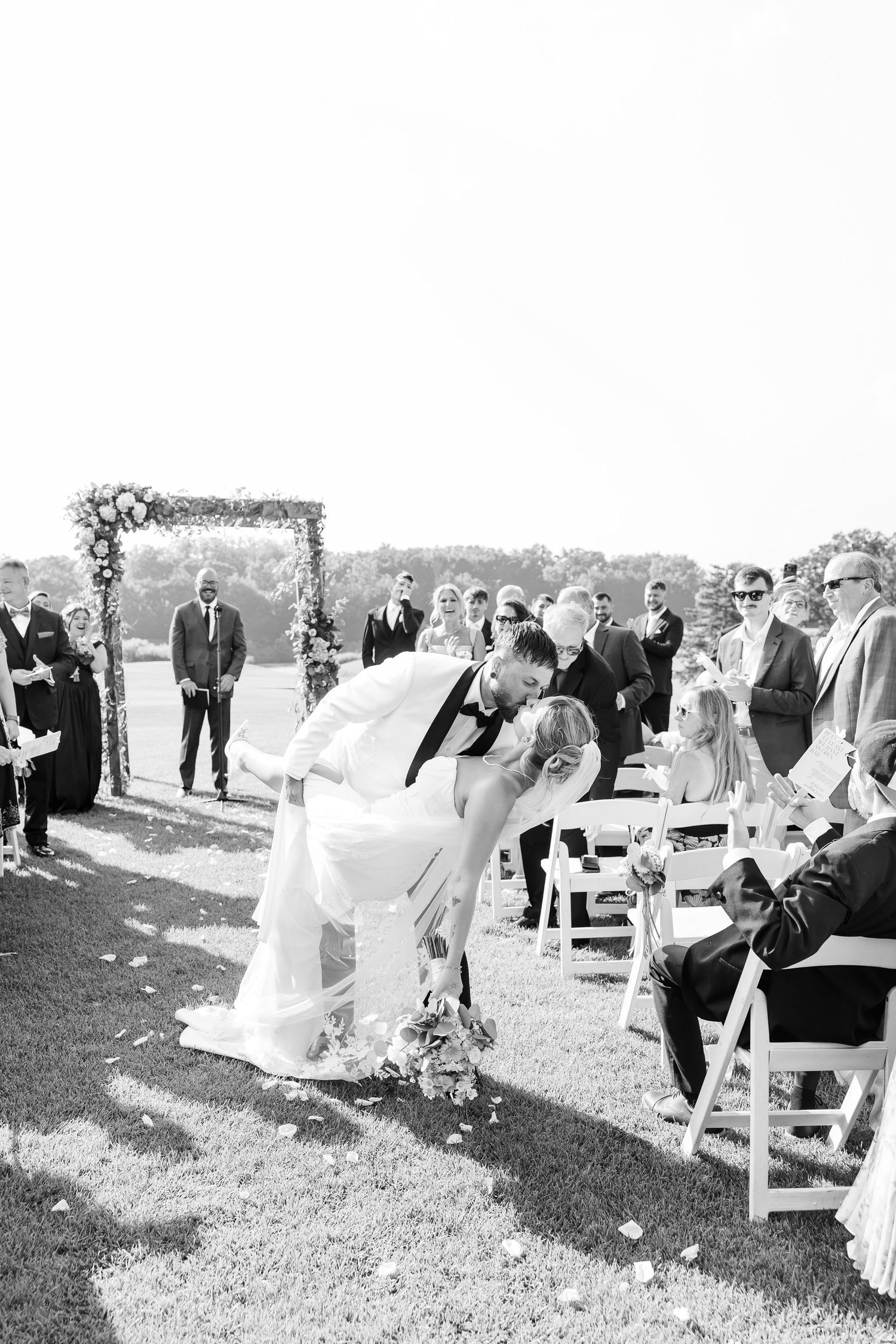 Black and white photo of bride and groom sharing a kiss at outdoor wedding ceremony with guests seated.