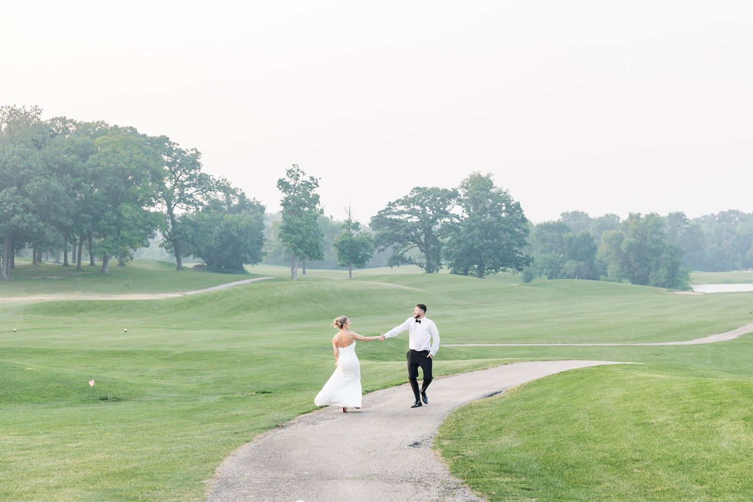 Bride and groom walking hand in hand along winding path on lush green golf course.