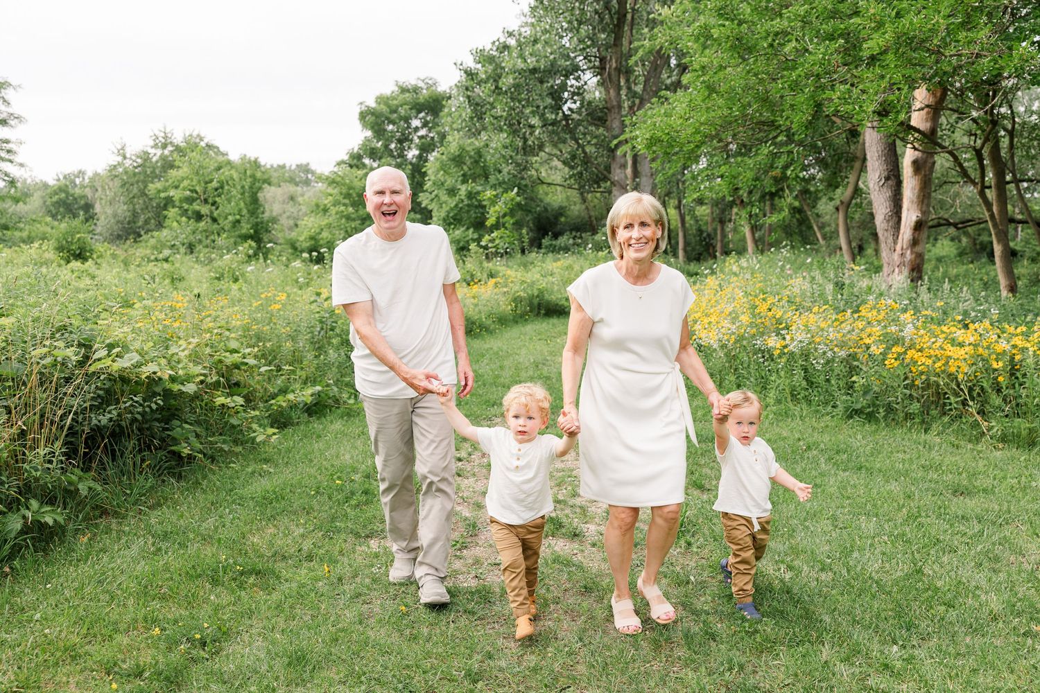 A joyful family of four holds hands while walking through a green meadow with yellow wildflowers.