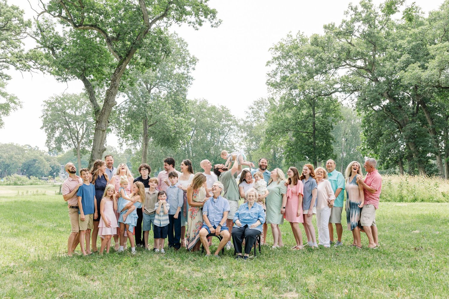 Large extended family posing together outdoors on green grass surrounded by tall trees on a sunny summer day.