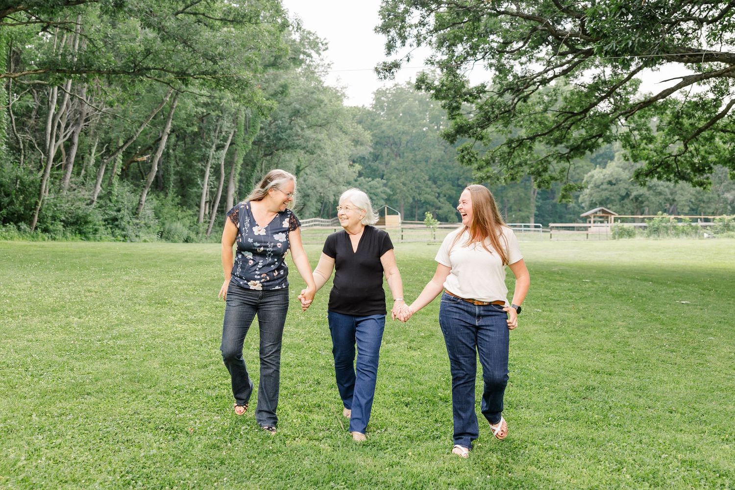 Three women holding hands and walking together through a lush green park surrounded by trees.