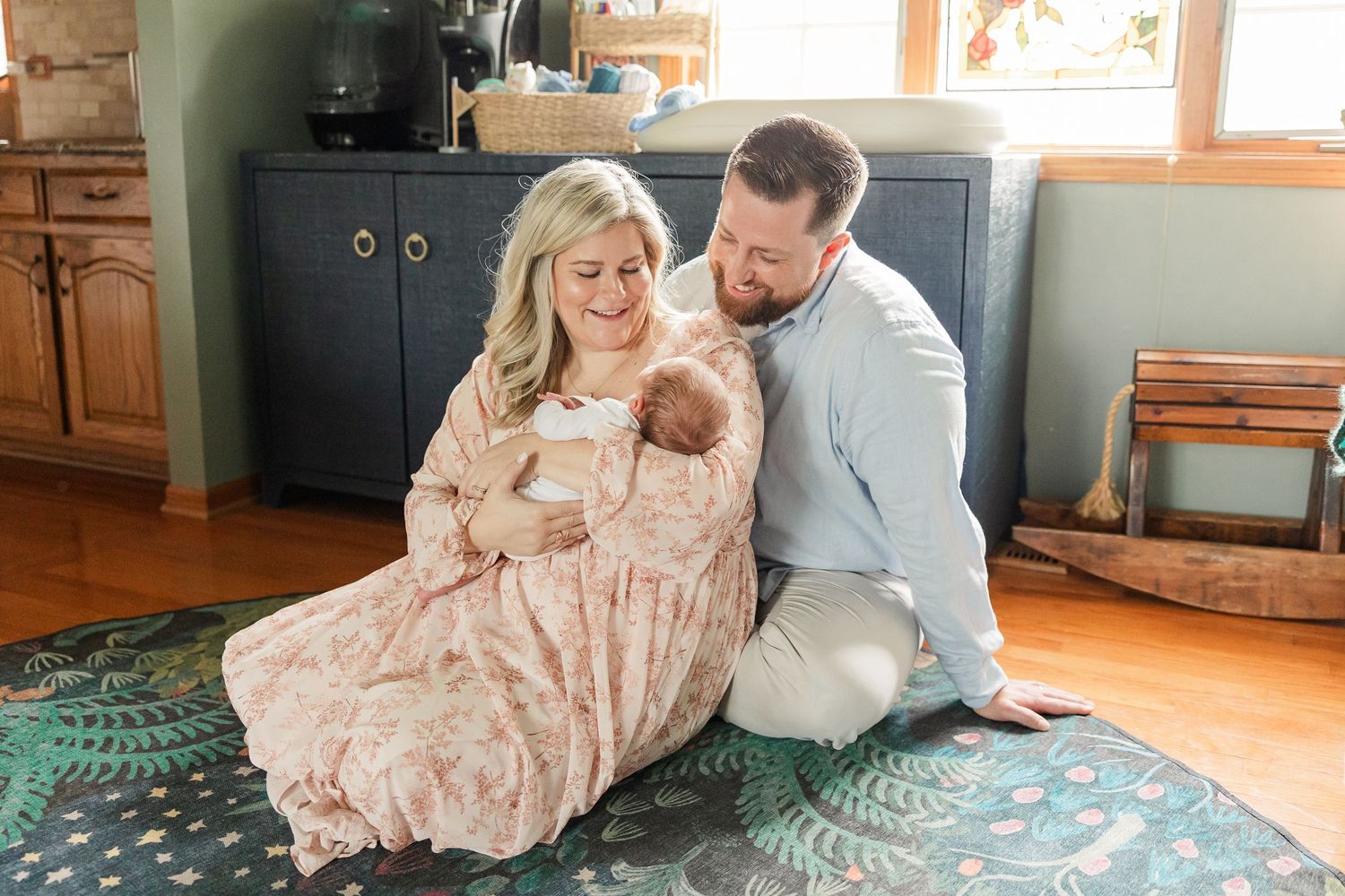 A couple sits on a rug holding their newborn baby, smiling lovingly in a warmly decorated home.