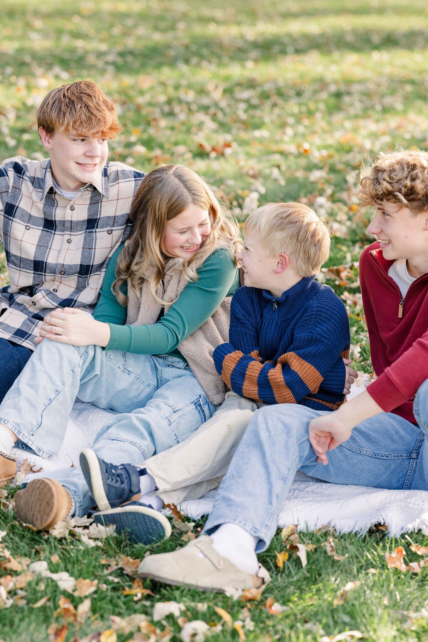 Three boys and a girl sit close together on a blanket among fallen autumn leaves, smiling and laughing during a family photo session.
