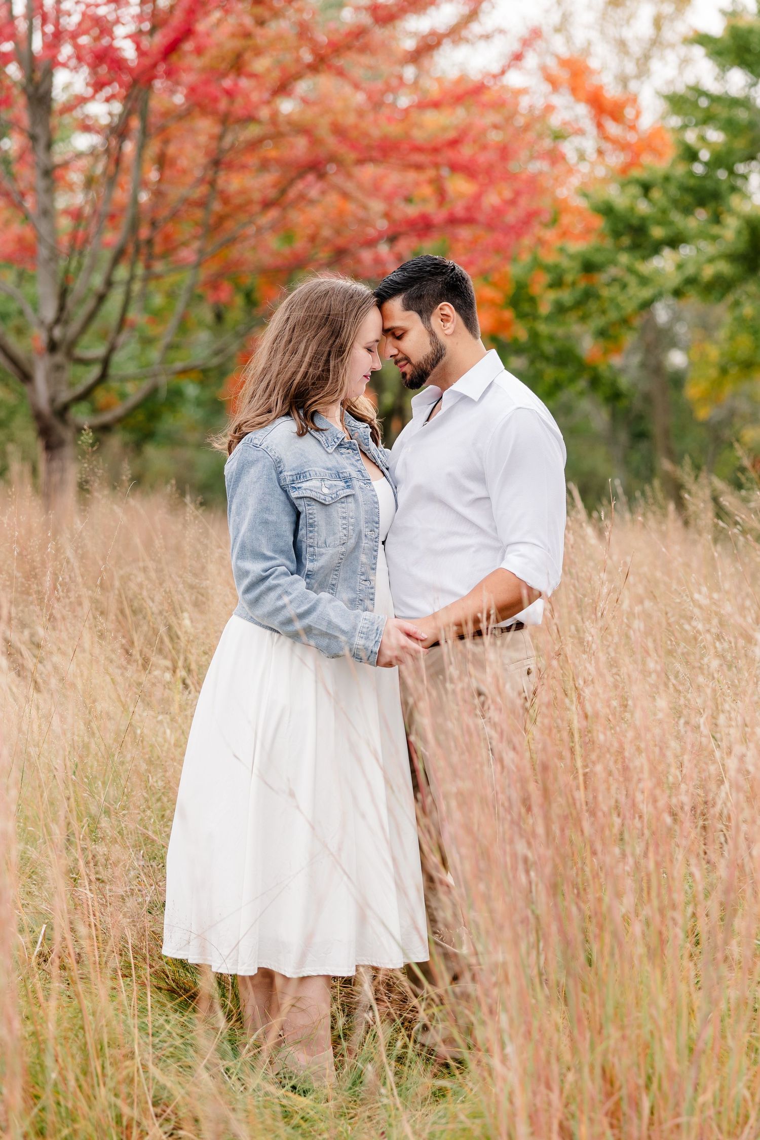 Engaged couple standing forehead to forehead in tall golden grass with colorful red autumn trees behind them.
