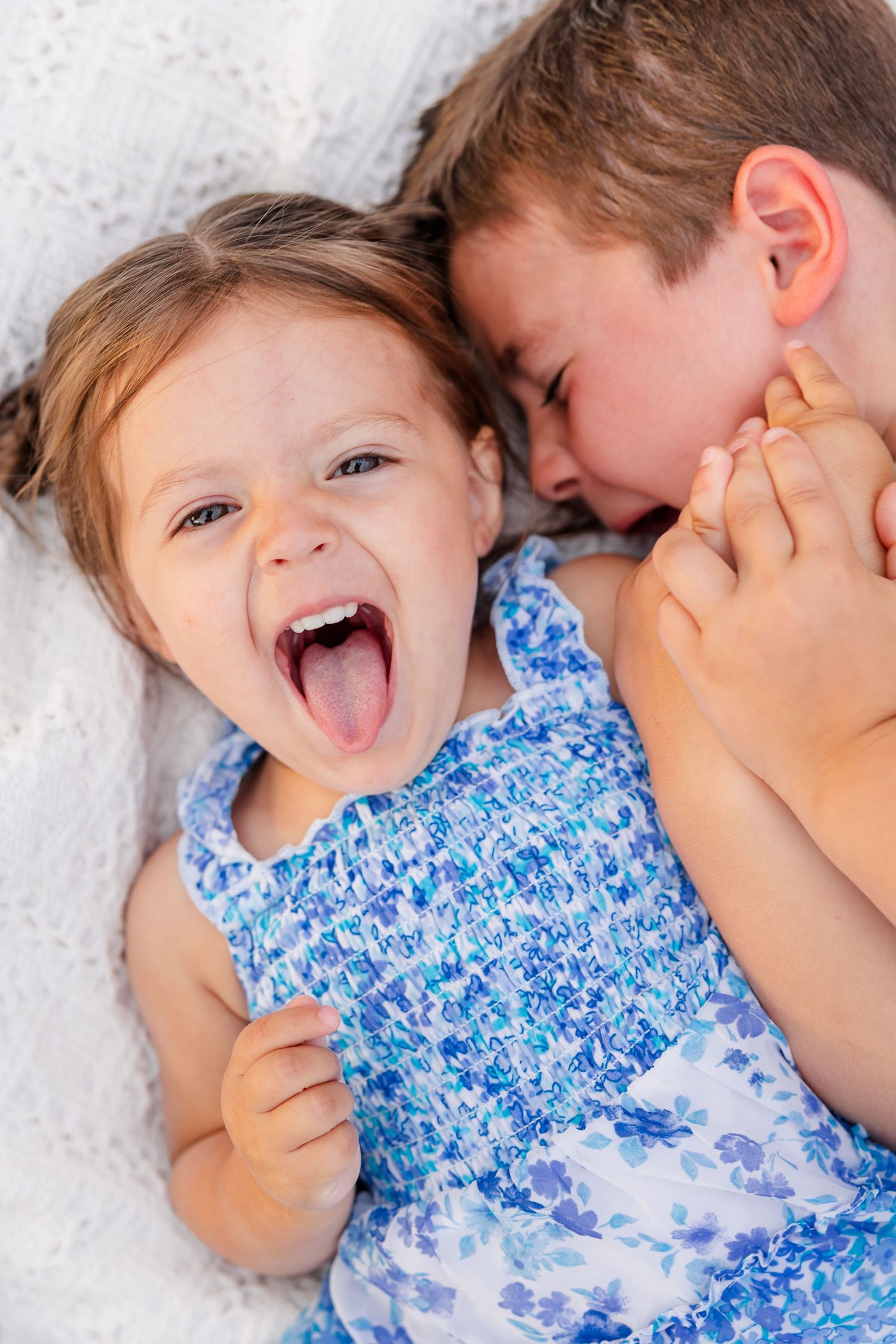 Laughing toddler girl in blue floral dress cuddles with older brother on white blanket.
