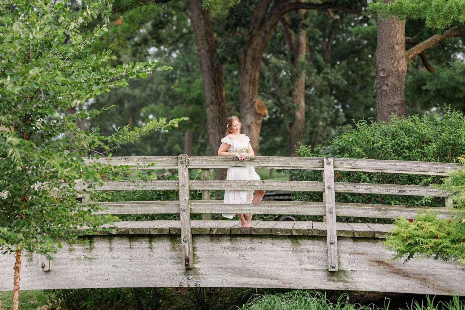 Wide senior portrait of Samantha standing on a wooden bridge surrounded by lush greenery at Cantigny Park.