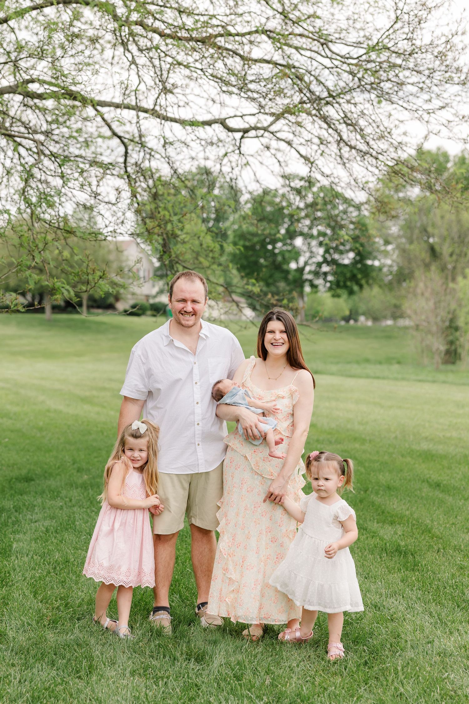 A happy family of five poses outdoors on green grass, parents with three young children including a newborn, dressed in soft pastel colors.