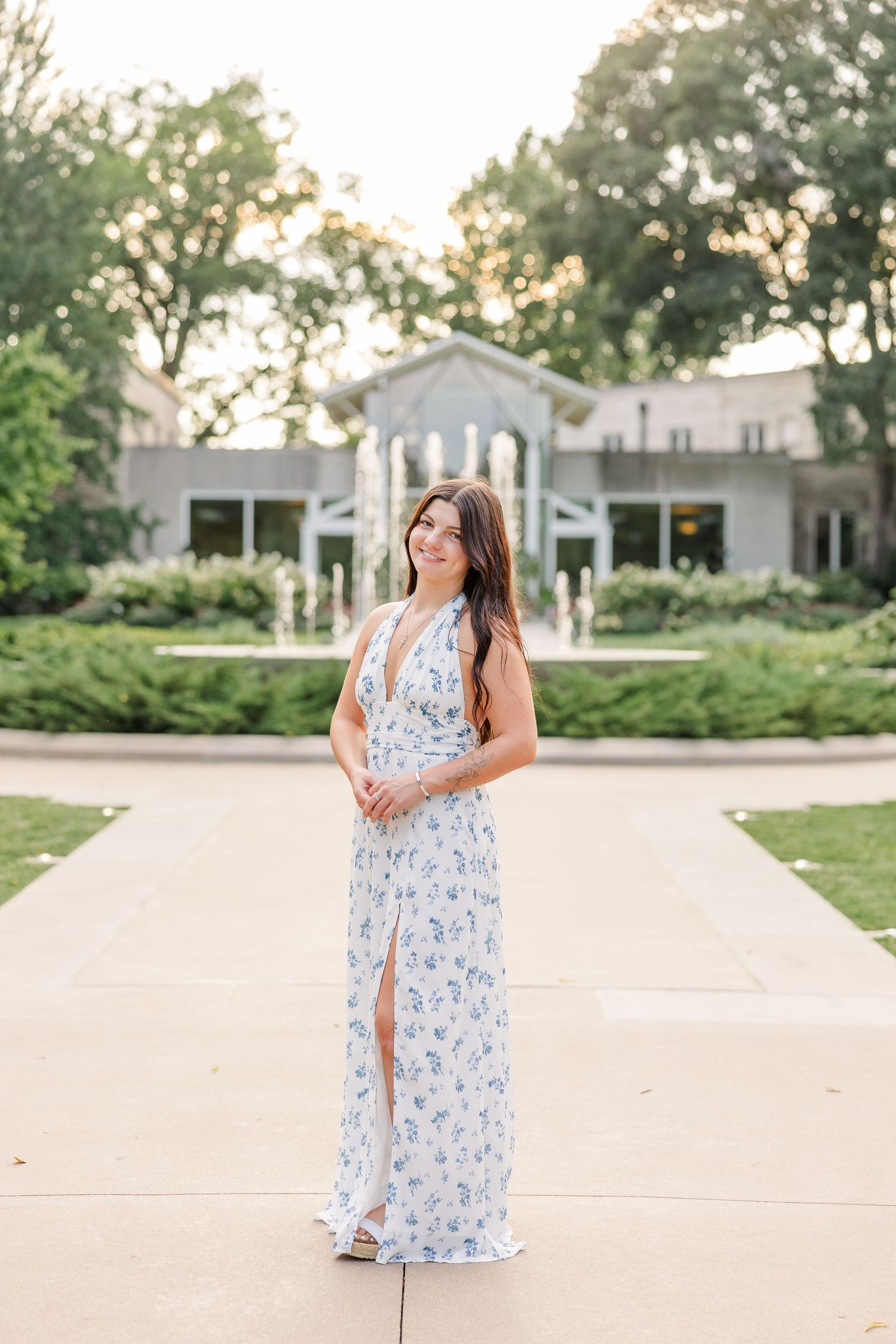 Brunette woman in white floral halter maxi dress stands on pathway before elegant fountain garden.