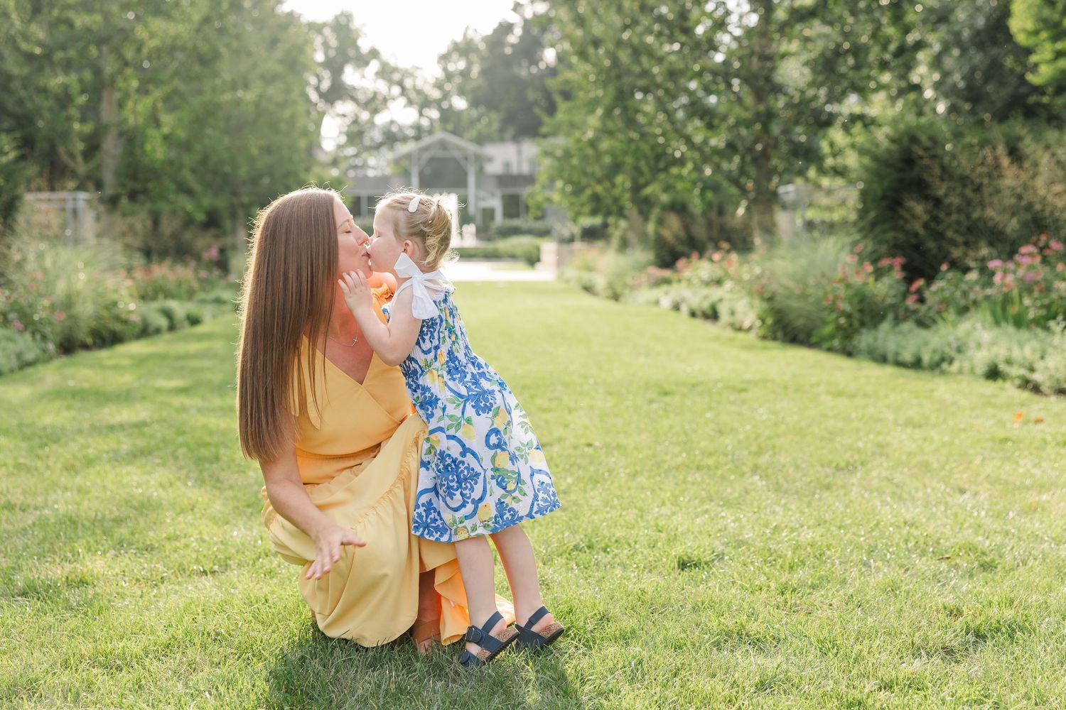 Mother crouching on green grass kissing her young daughter in a floral dress in a beautiful garden.