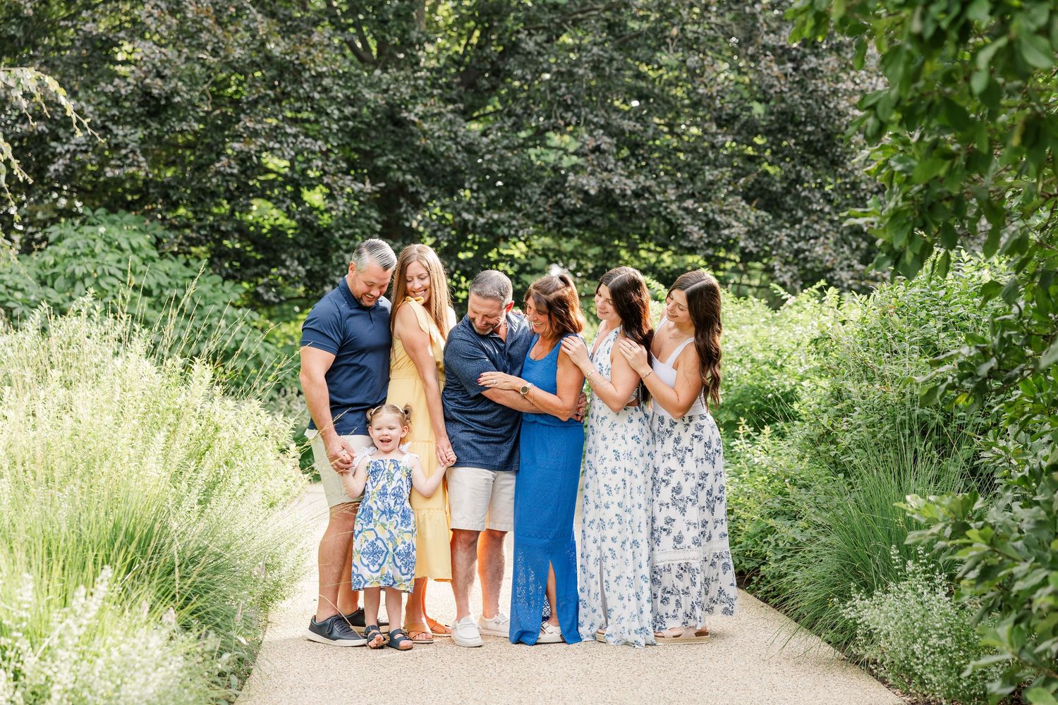 Family group hugging on a garden path surrounded by lush green trees and plants on a sunny day.