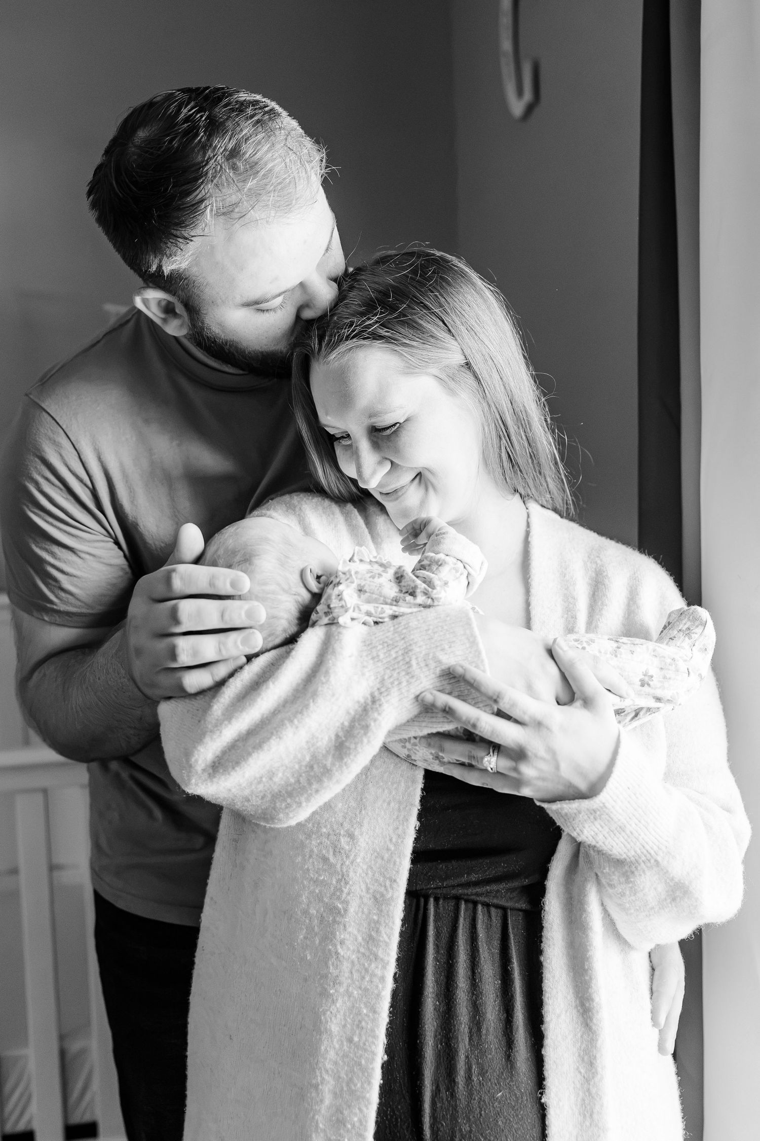 Black and white photo of a bearded father kissing mothers head as they both lovingly hold their newborn.