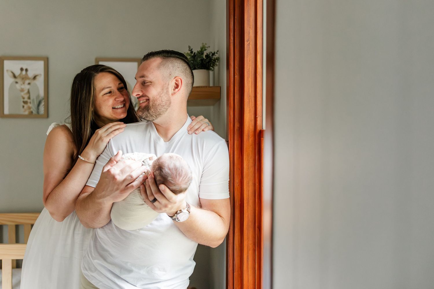 Happy couple in white outfits holding their newborn baby, smiling together in a bright nursery room.
