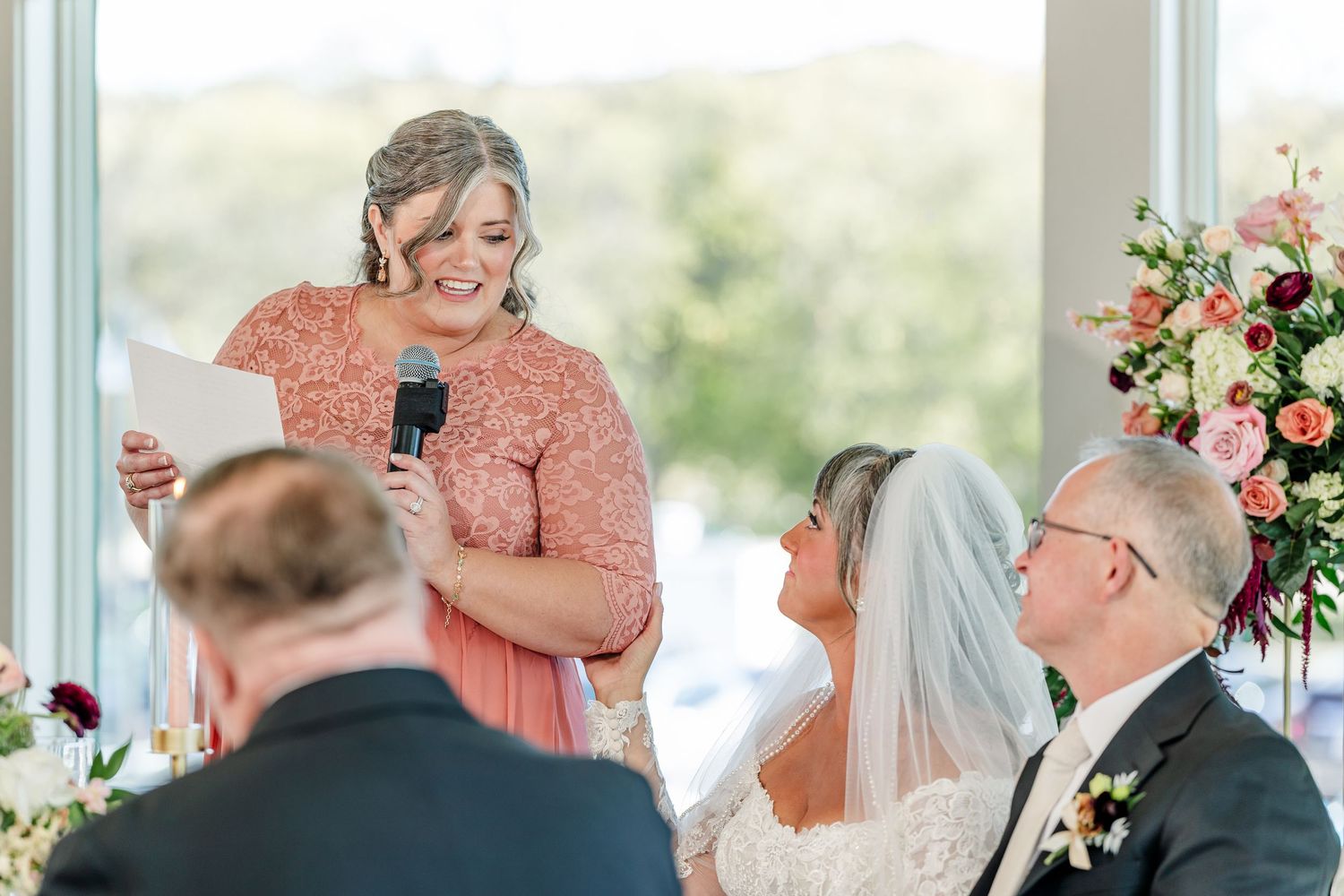 Woman in a coral lace dress gives a wedding toast into a microphone as the bride in a veil and groom smile beside floral arrangements.