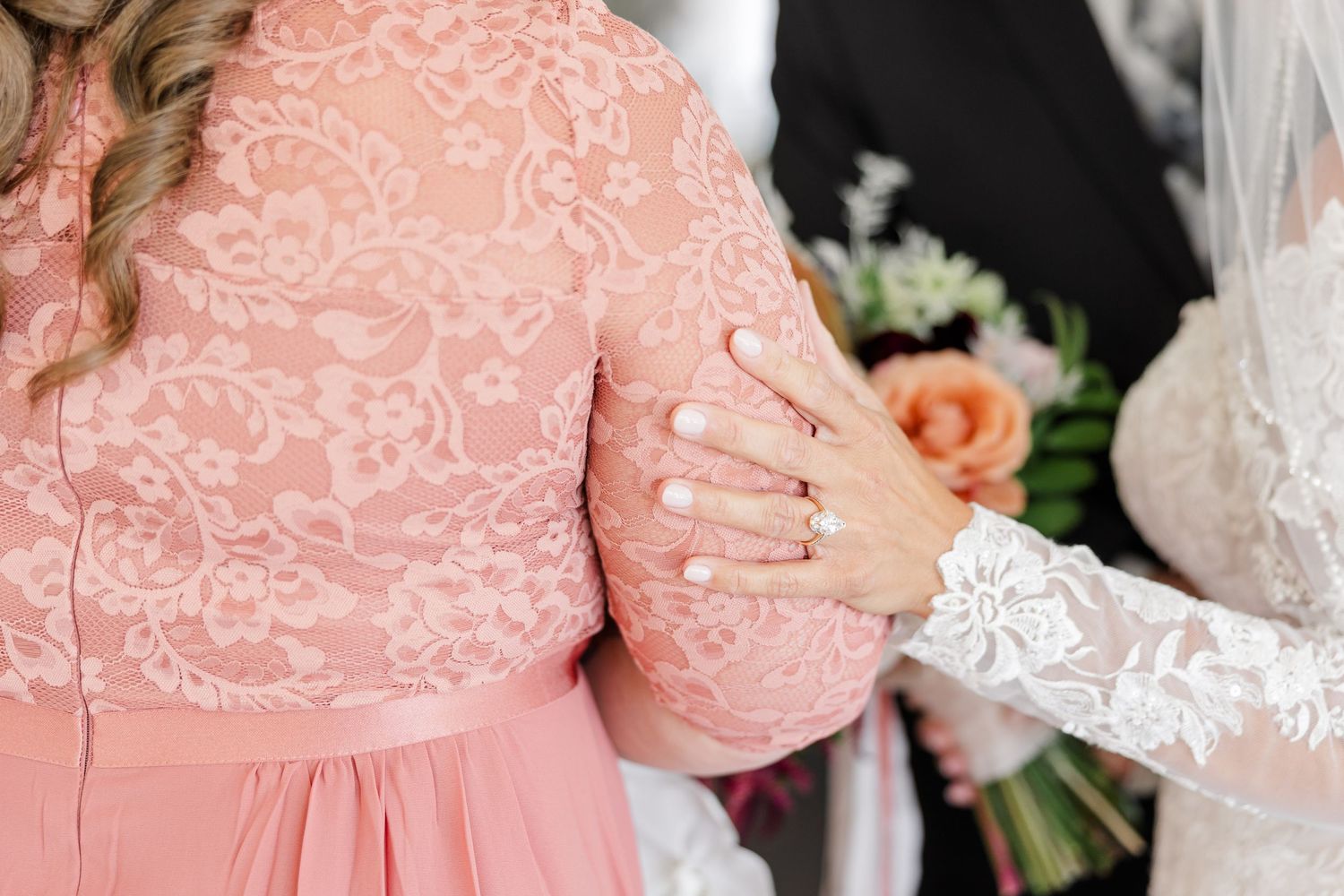 Bride in a white lace sleeve rests her hand on the arm of a guest wearing a blush pink lace dress, with a floral bouquet nearby.