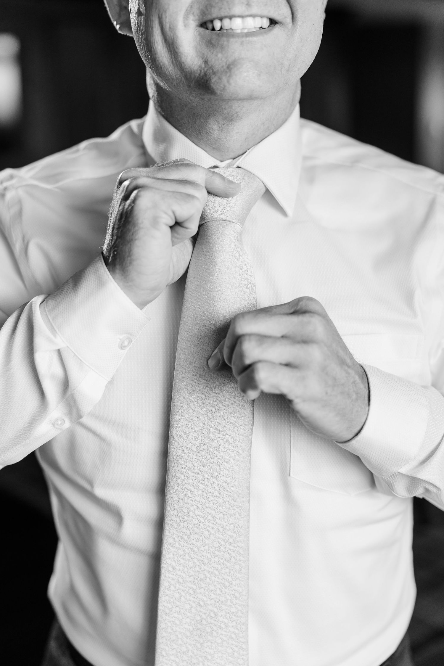 Smiling man in a white dress shirt adjusts his patterned necktie while getting dressed, shown in black and white.