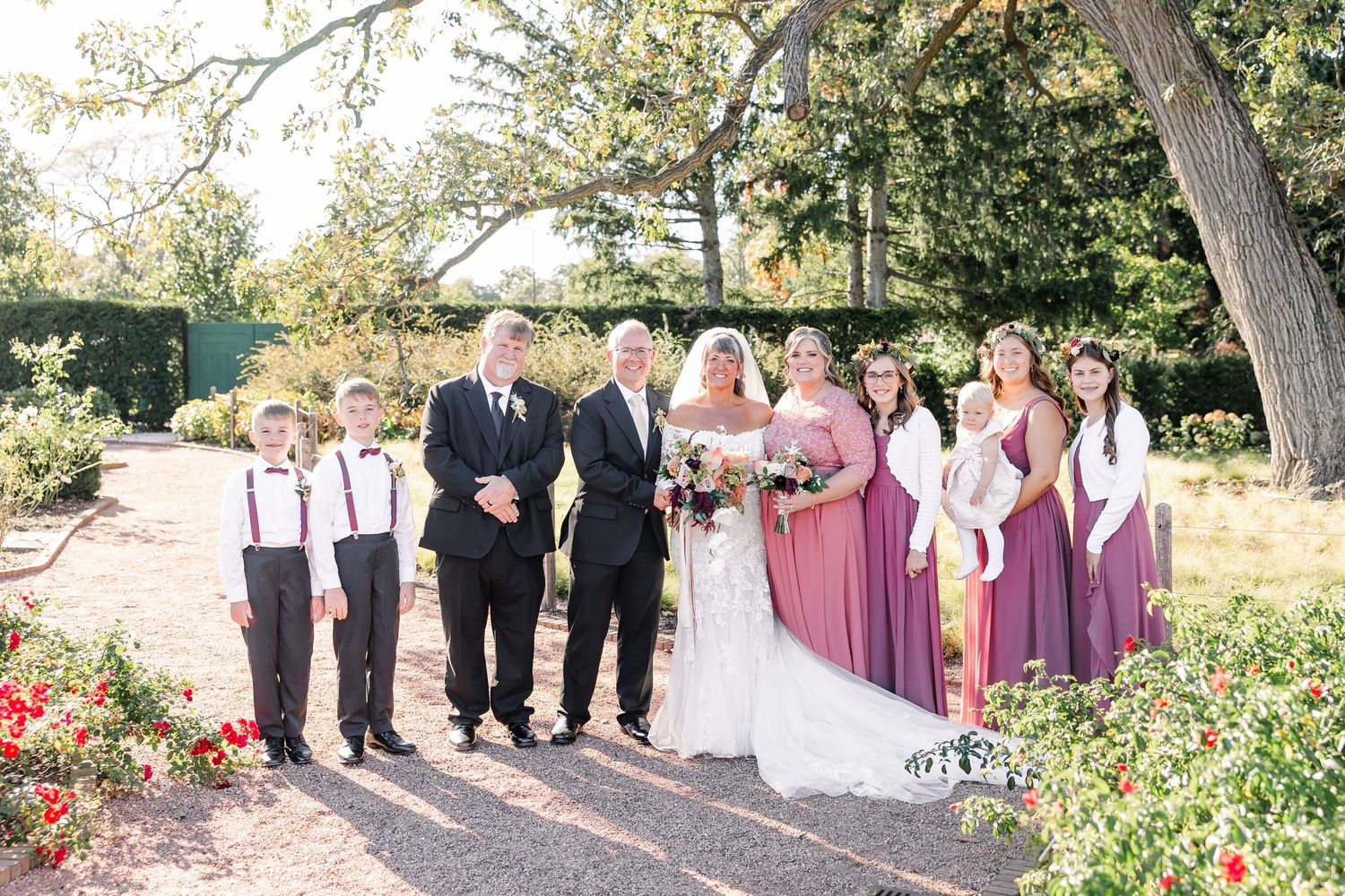 Bride and groom pose outdoors with groomsmen, young ring bearers and bridesmaids in mauve dresses in a sunlit garden wedding party portrait.