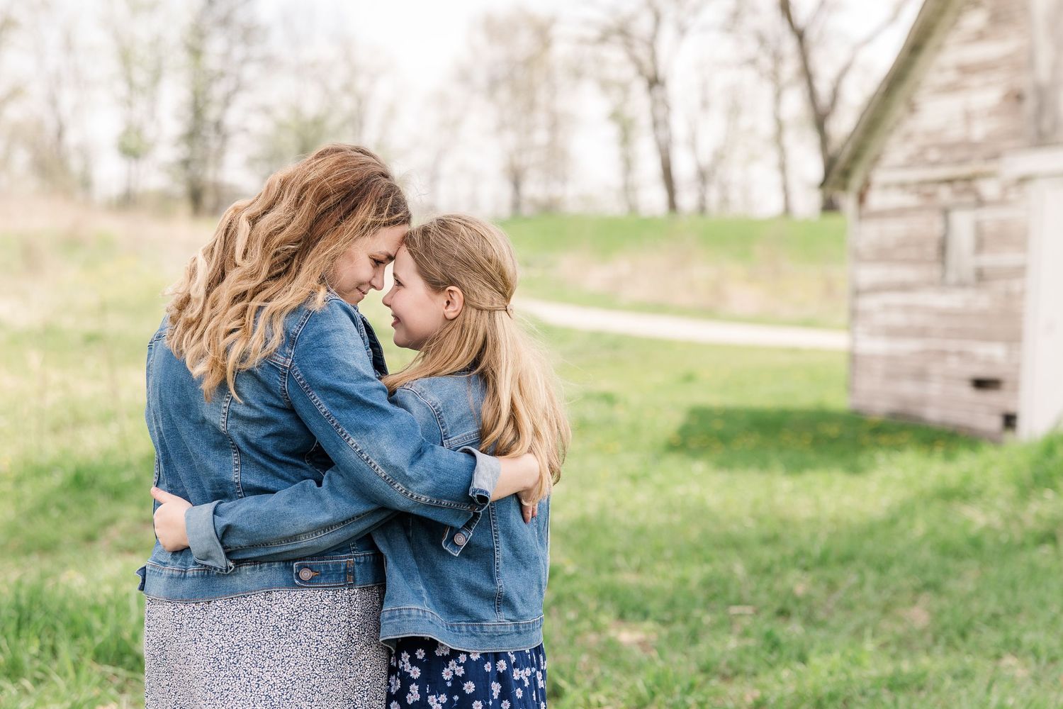 Two people share an intimate embrace while wearing denim in a rustic outdoor setting with a wooden structure.