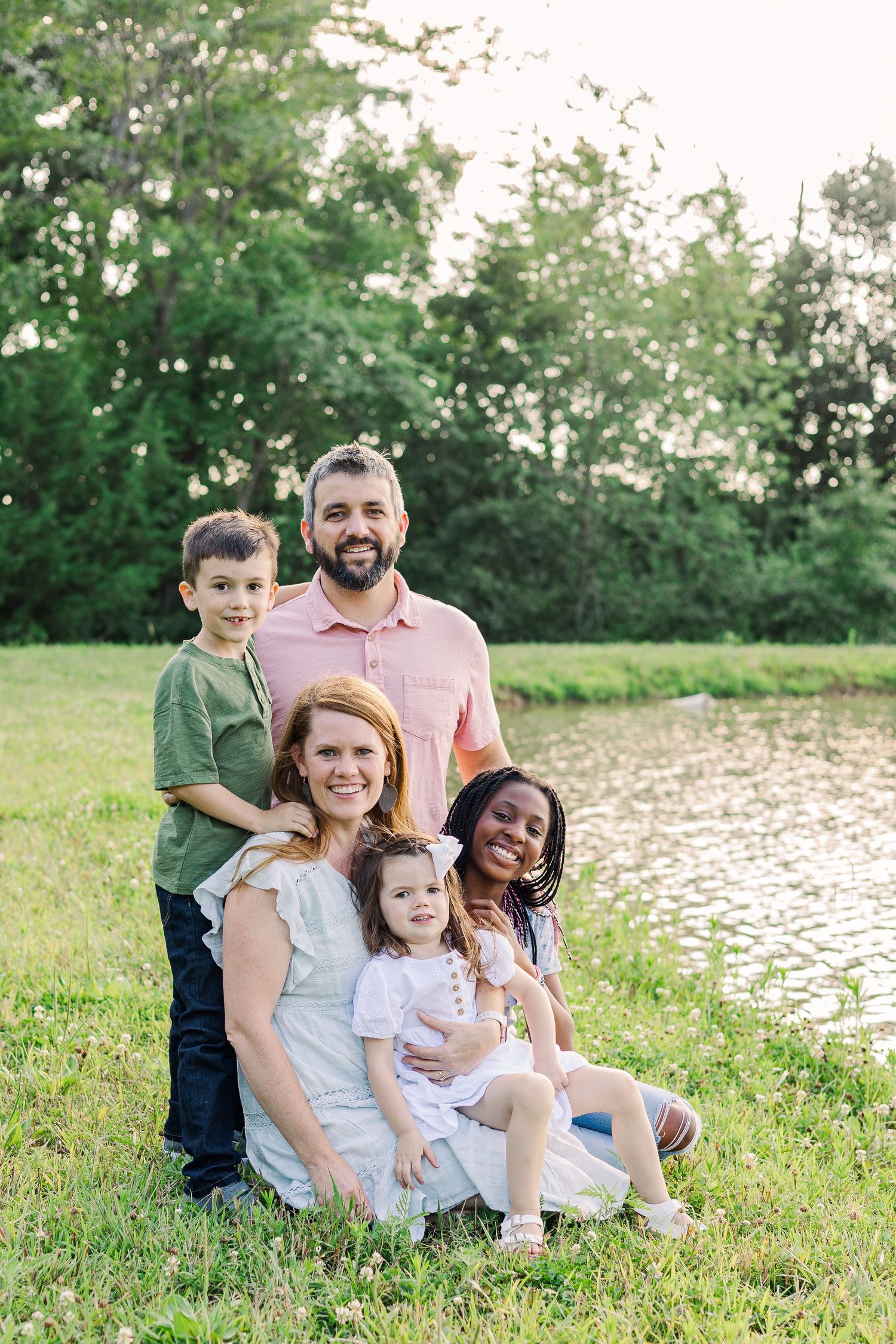A happy family group sits together on grassy hill beside a pond on a sunny day.