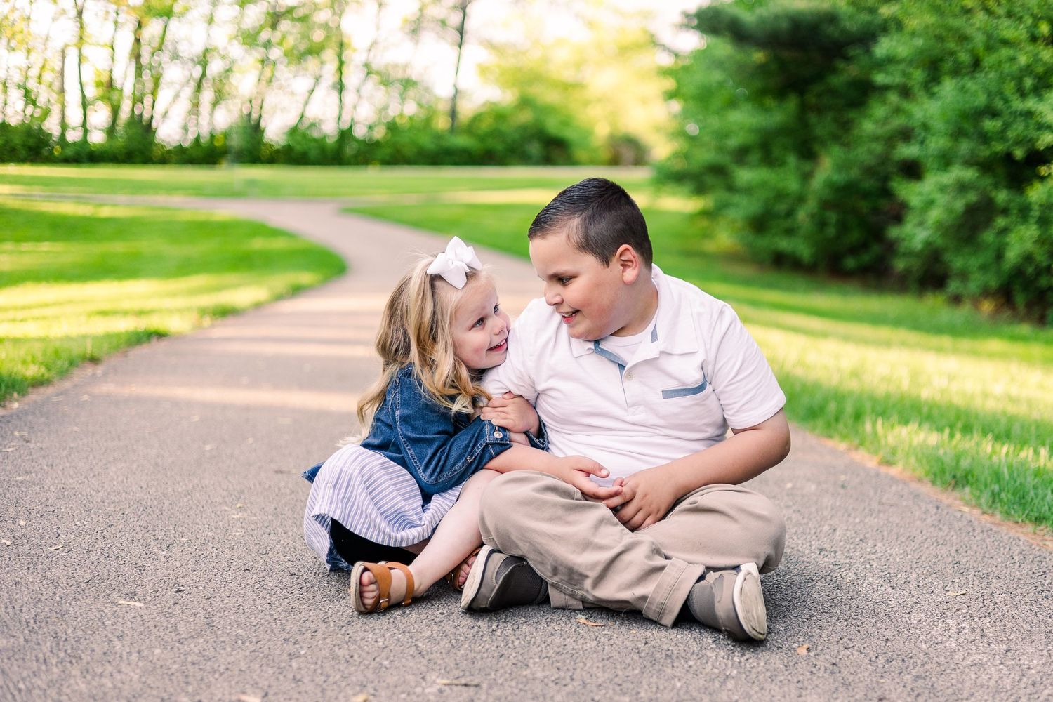 Two people share a laugh while sitting on a paved path in a sunny park setting.