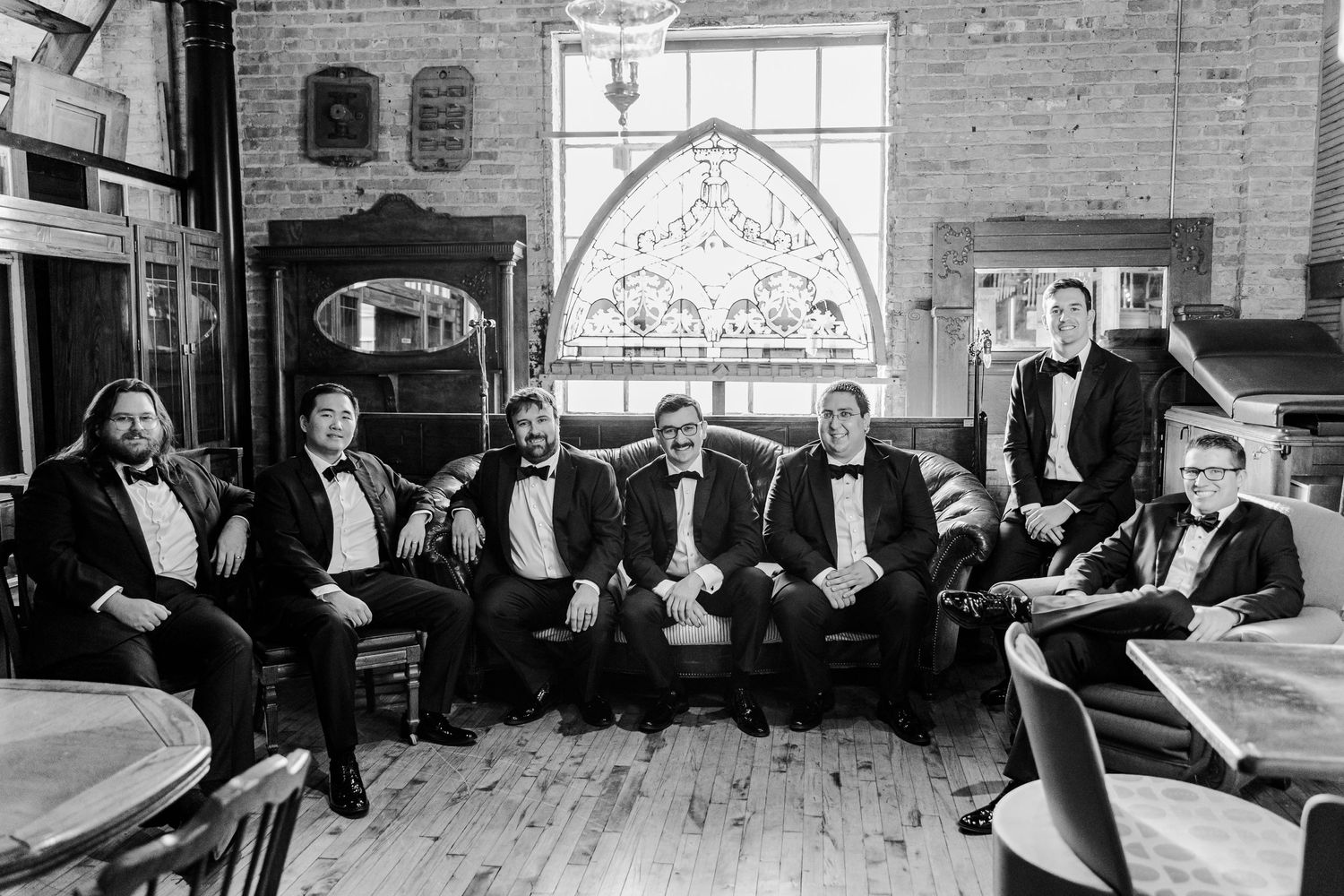 Black and white photo of groomsmen relaxing in formal attire in historic brick-walled room.
