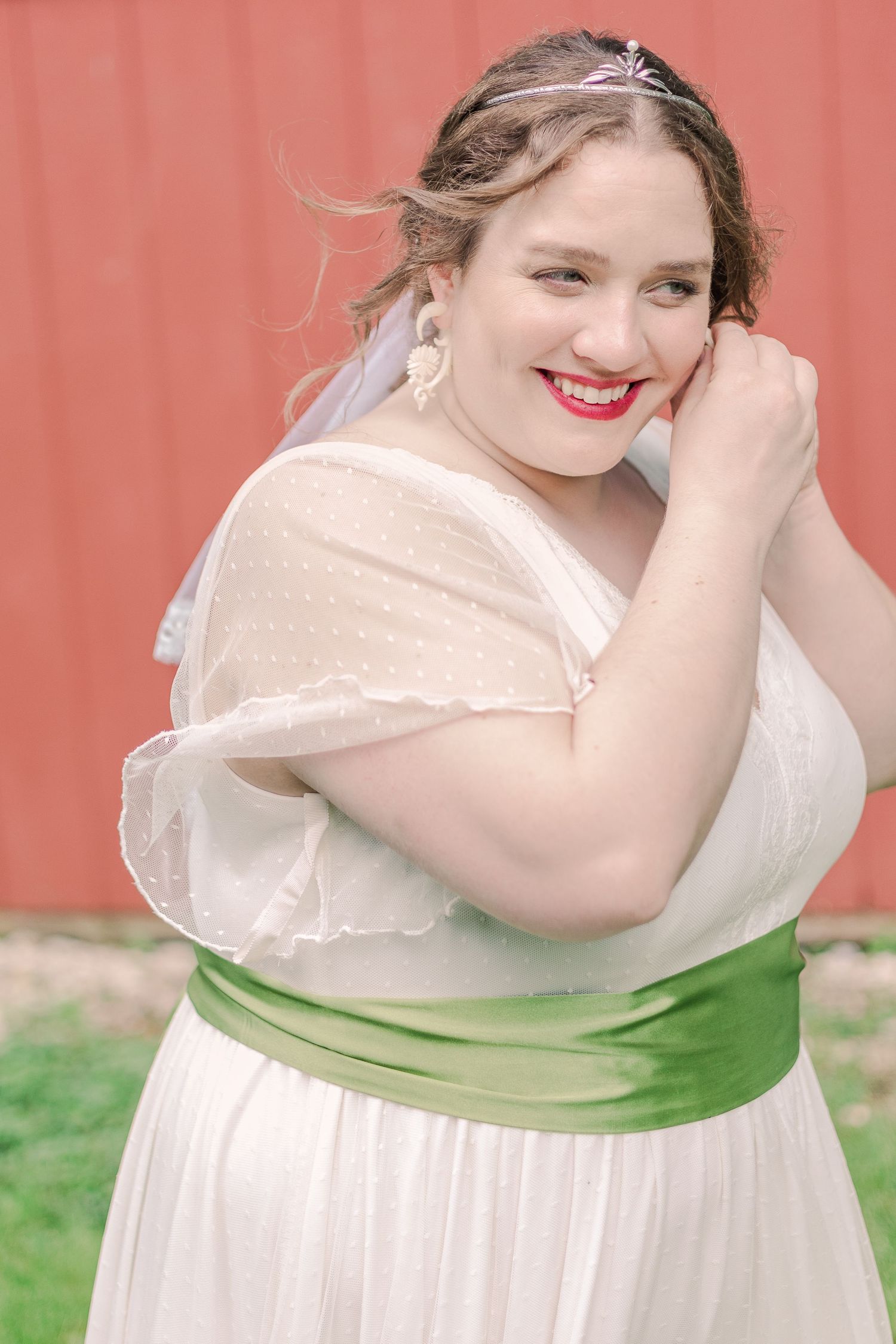Person in wedding dress with green sash poses against red barn background.