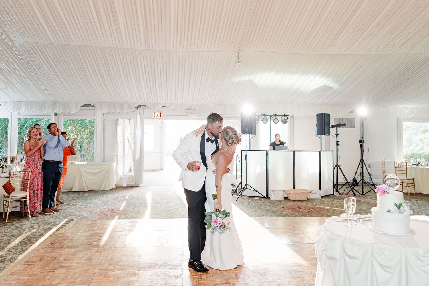 Bride and groom share first dance in elegant white tent reception venue with DJ and guests watching.