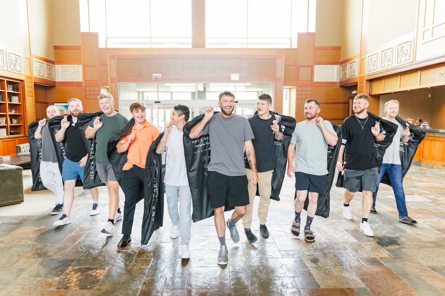 Group of groomsmen laughing and posing together in a bright indoor venue before a wedding.
