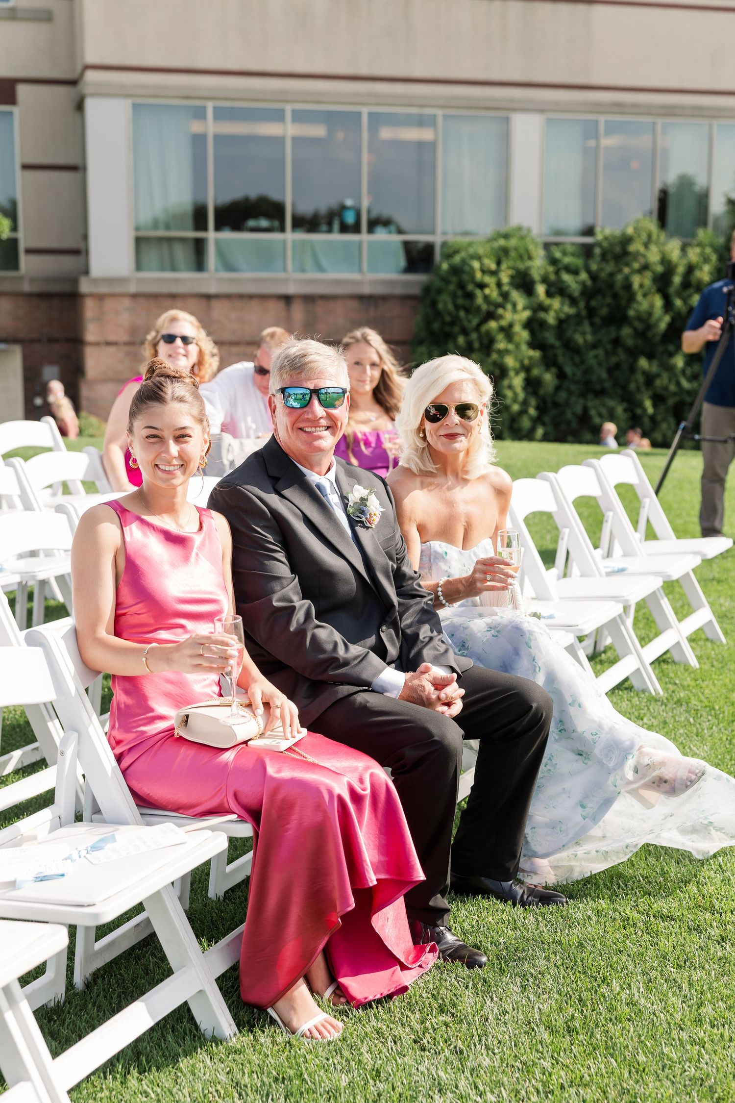 Wedding guests seated in white chairs on a lawn, smiling at an outdoor ceremony on a sunny day.
