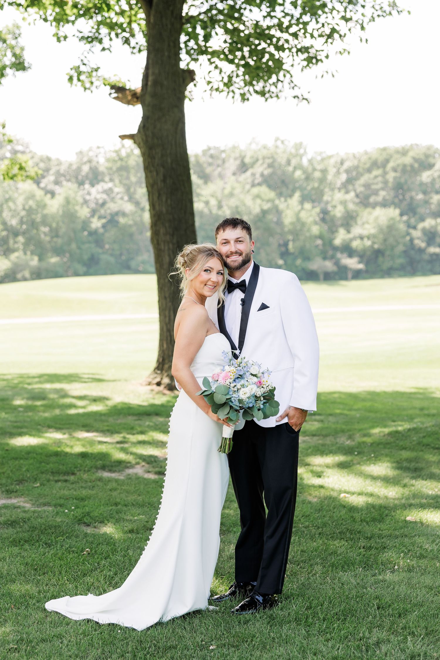 Bride in white gown and groom in white tuxedo posing together outdoors on green lawn.