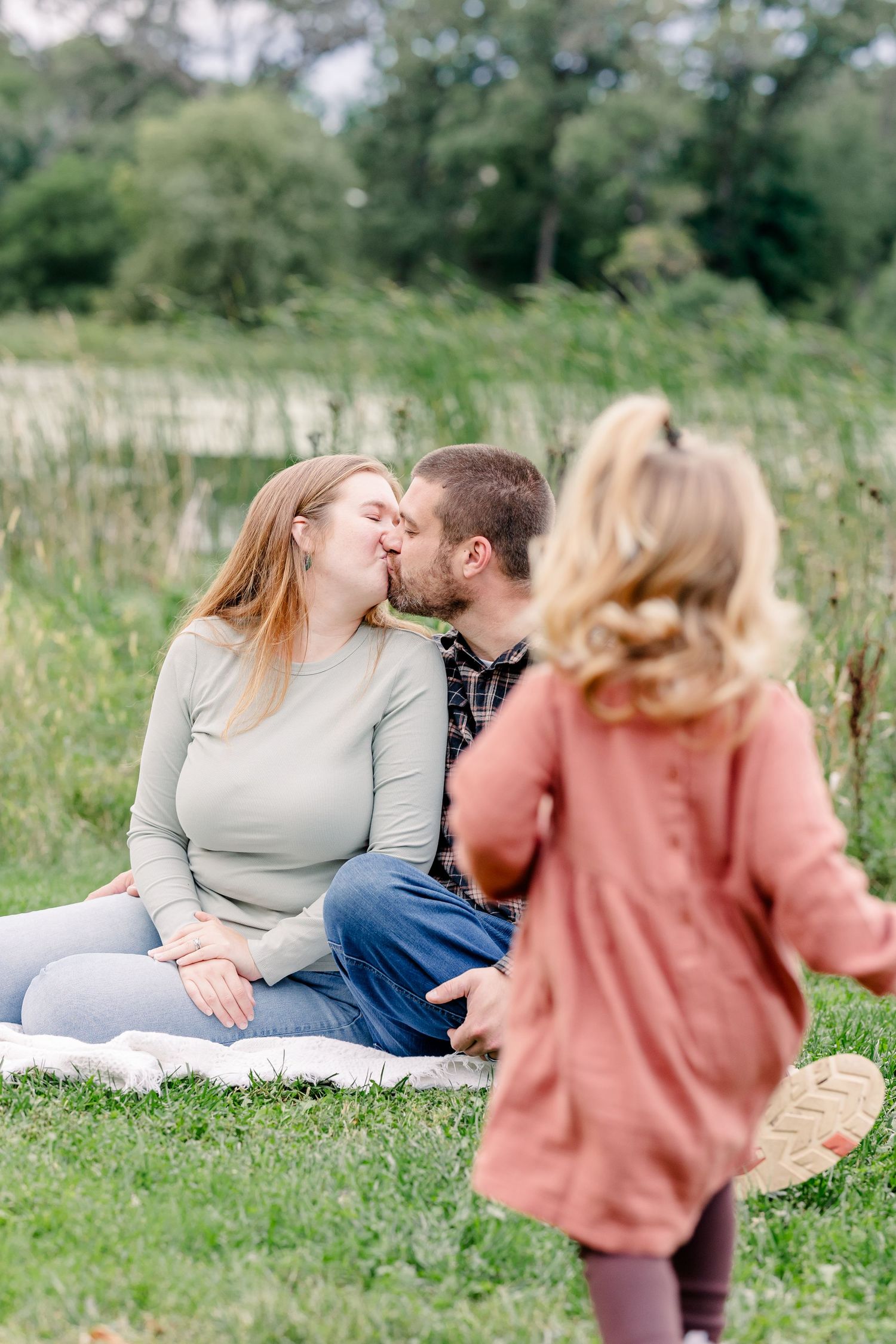 A couple shares a kiss on a blanket in a green meadow as their young daughter runs toward them.