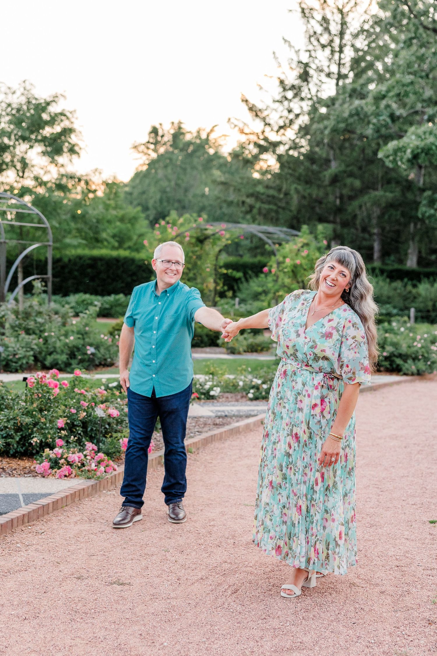 Smiling couple holding hands in rose garden, woman in floral dress, man in teal shirt at sunset.