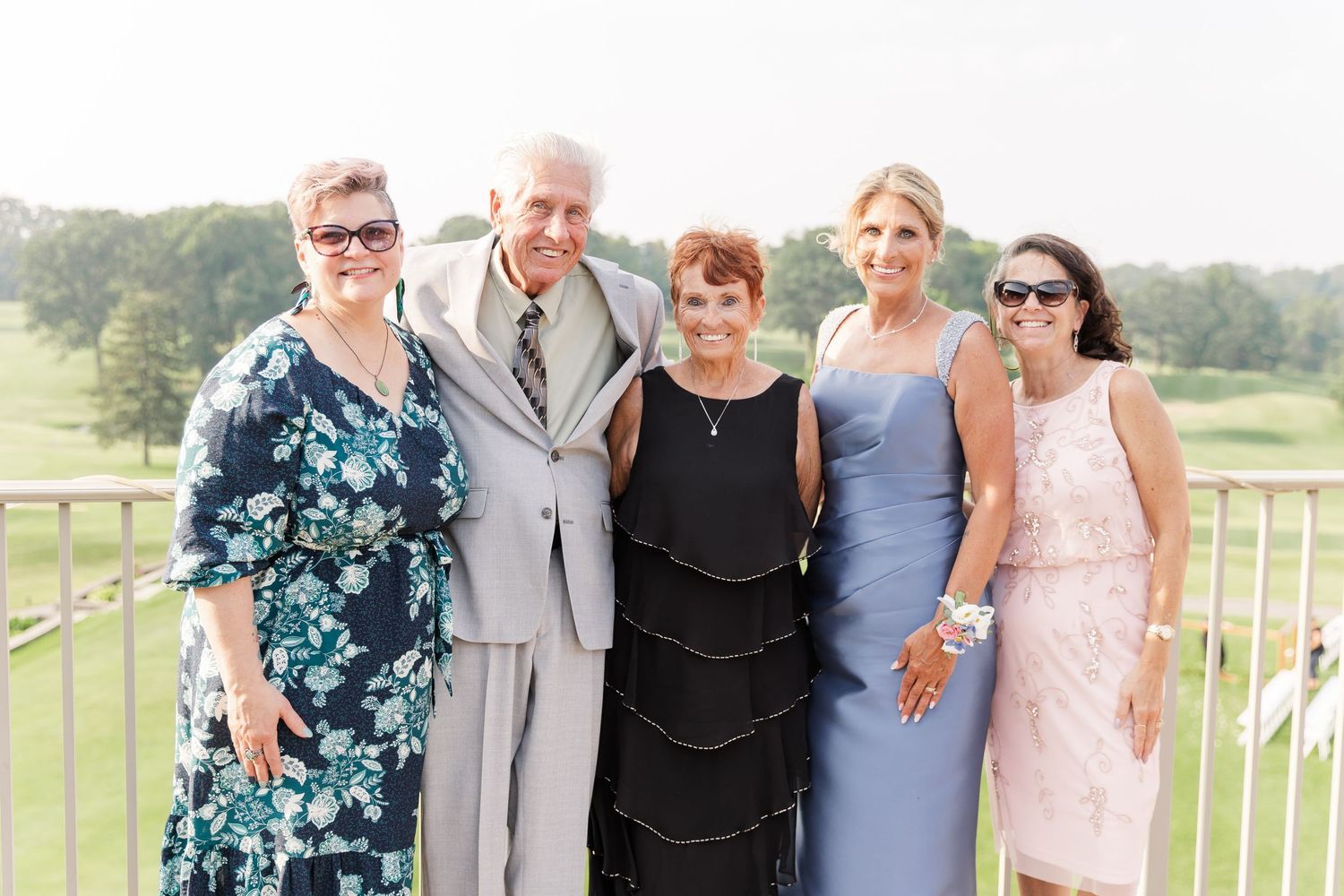 Five wedding guests smile together on an outdoor terrace overlooking a golf course at a summer wedding.
