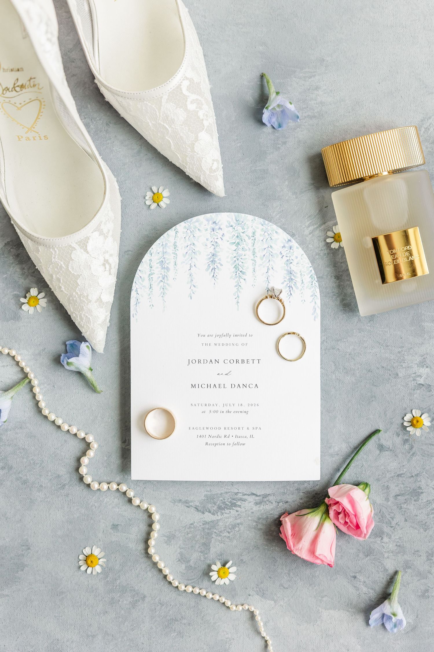 Wedding flat lay featuring gold rings on an arch invitation with lace heels, pearl necklace, perfume, and fresh flowers on gray.