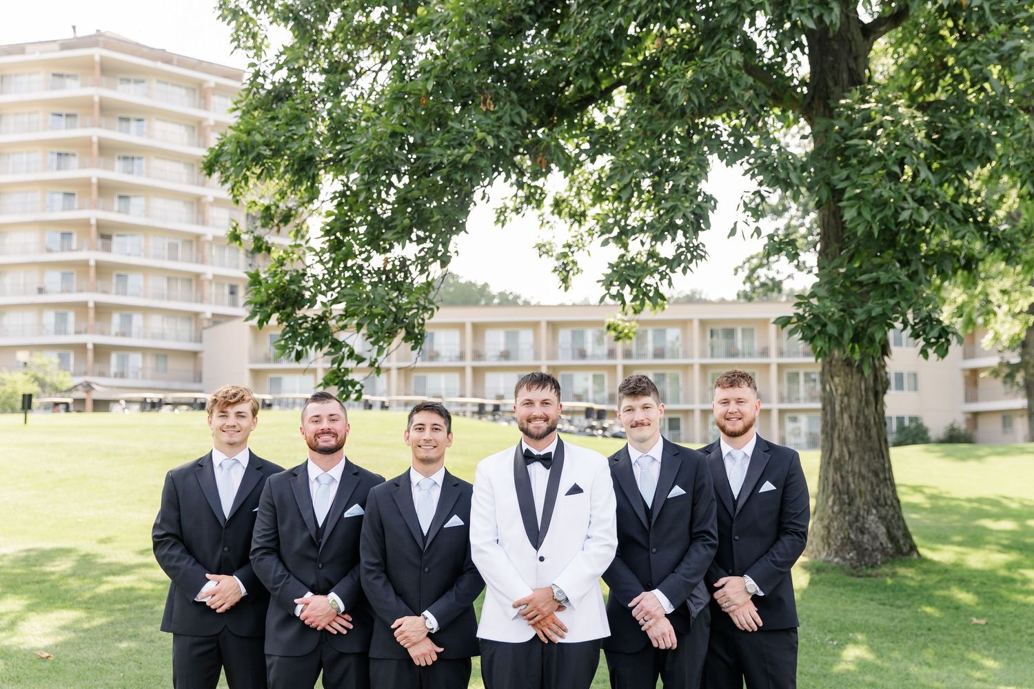 Groom in white tuxedo jacket stands with five groomsmen in black suits outdoors on wedding day.