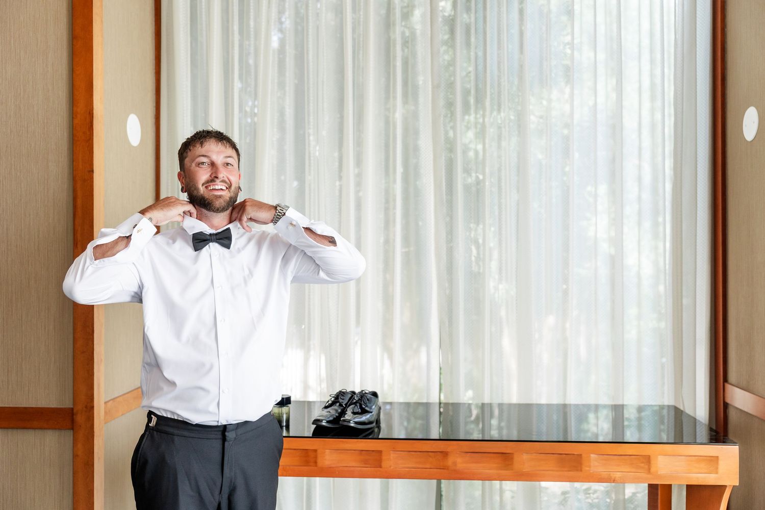 Smiling groom in white shirt and black bow tie adjusting his collar before wedding ceremony.