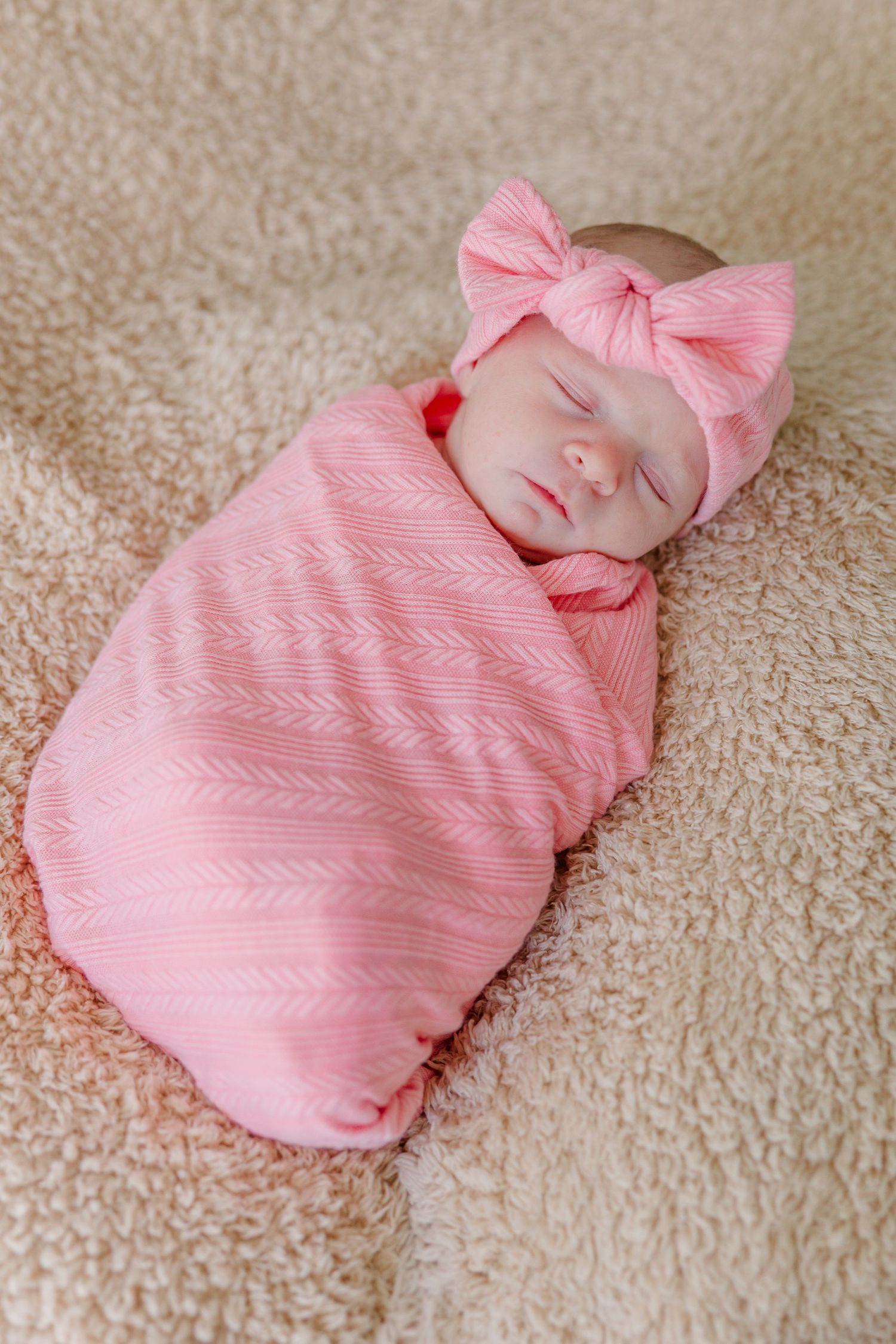 Sleeping newborn baby girl swaddled in pink wrap with matching bow headband on soft beige blanket.