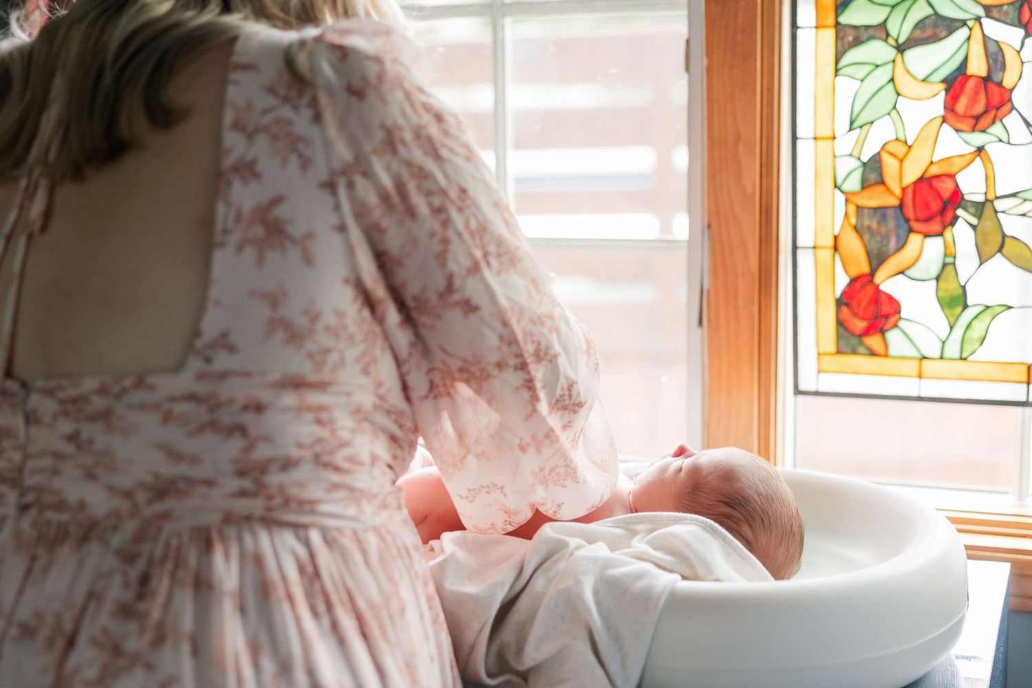 Mother in floral dress tenderly holding newborn baby near stained glass window with rose design.