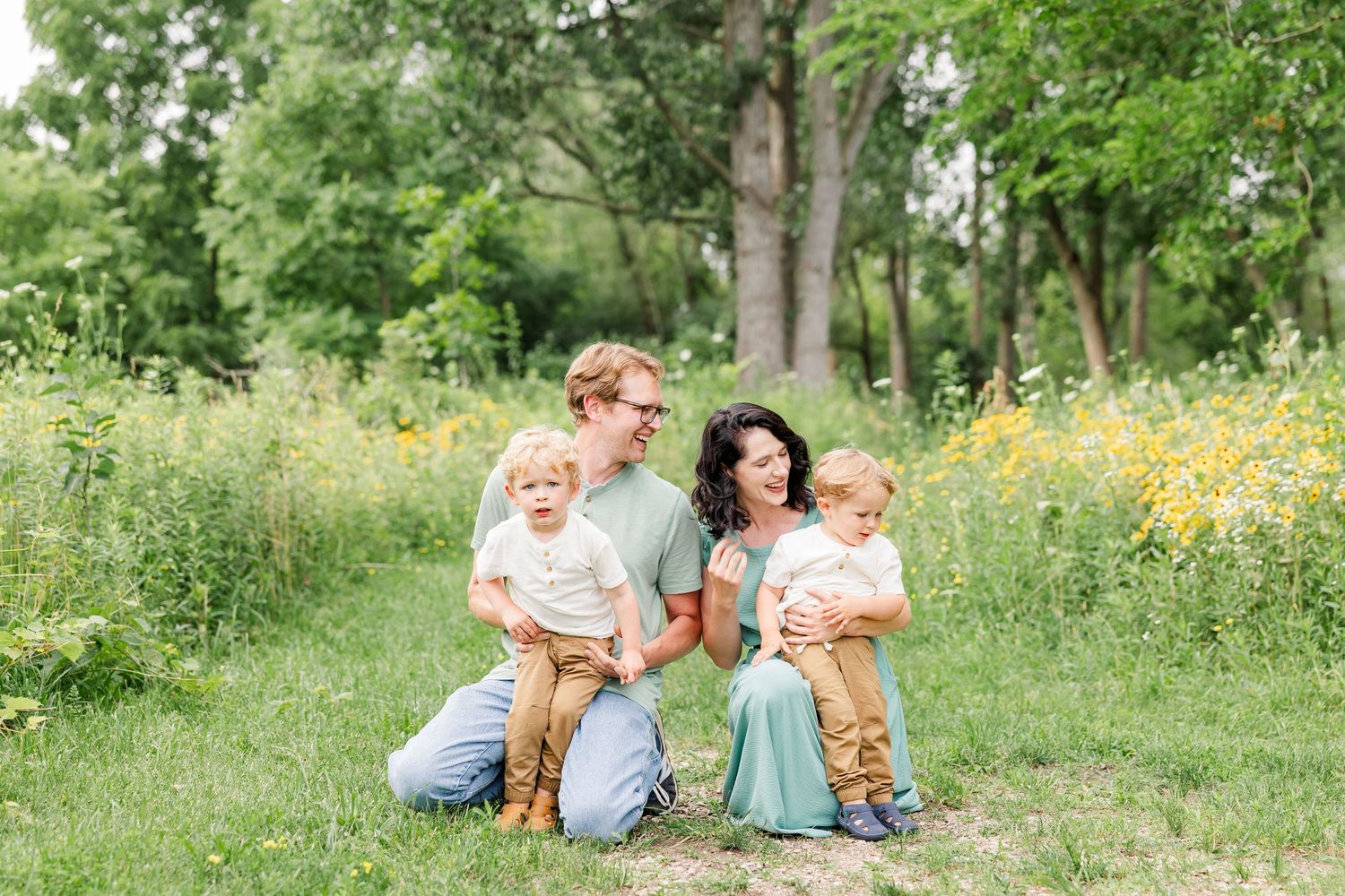A young family of four sits together in a sunny wildflower meadow surrounded by lush green trees.