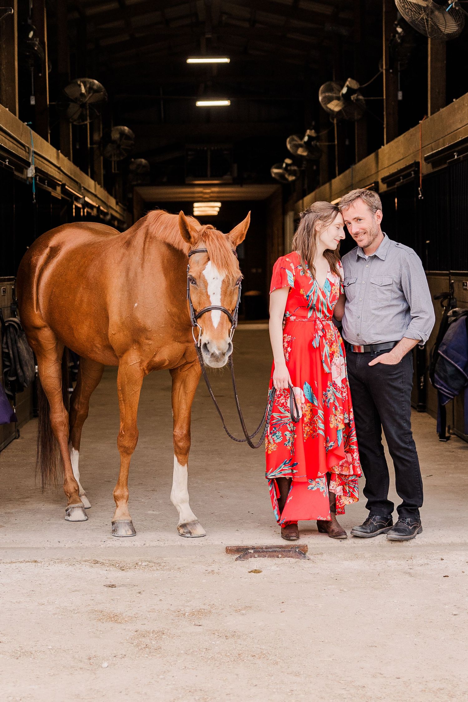 A beautiful chestnut horse poses with a couple in a rustic barn setting.