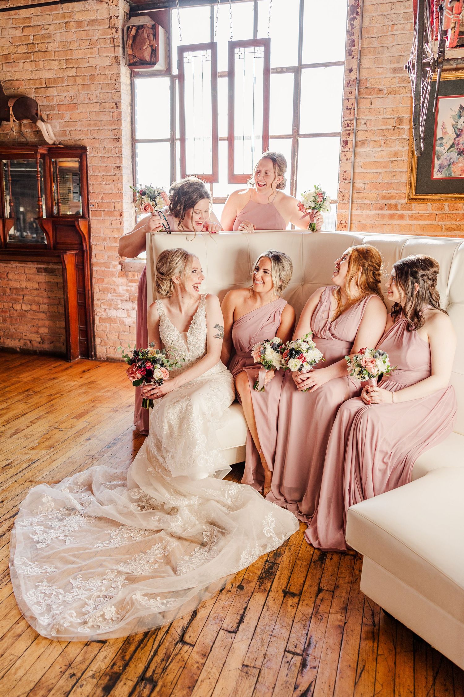 Wedding party lounges on cream couch in rustic brick venue with natural light streaming through windows.