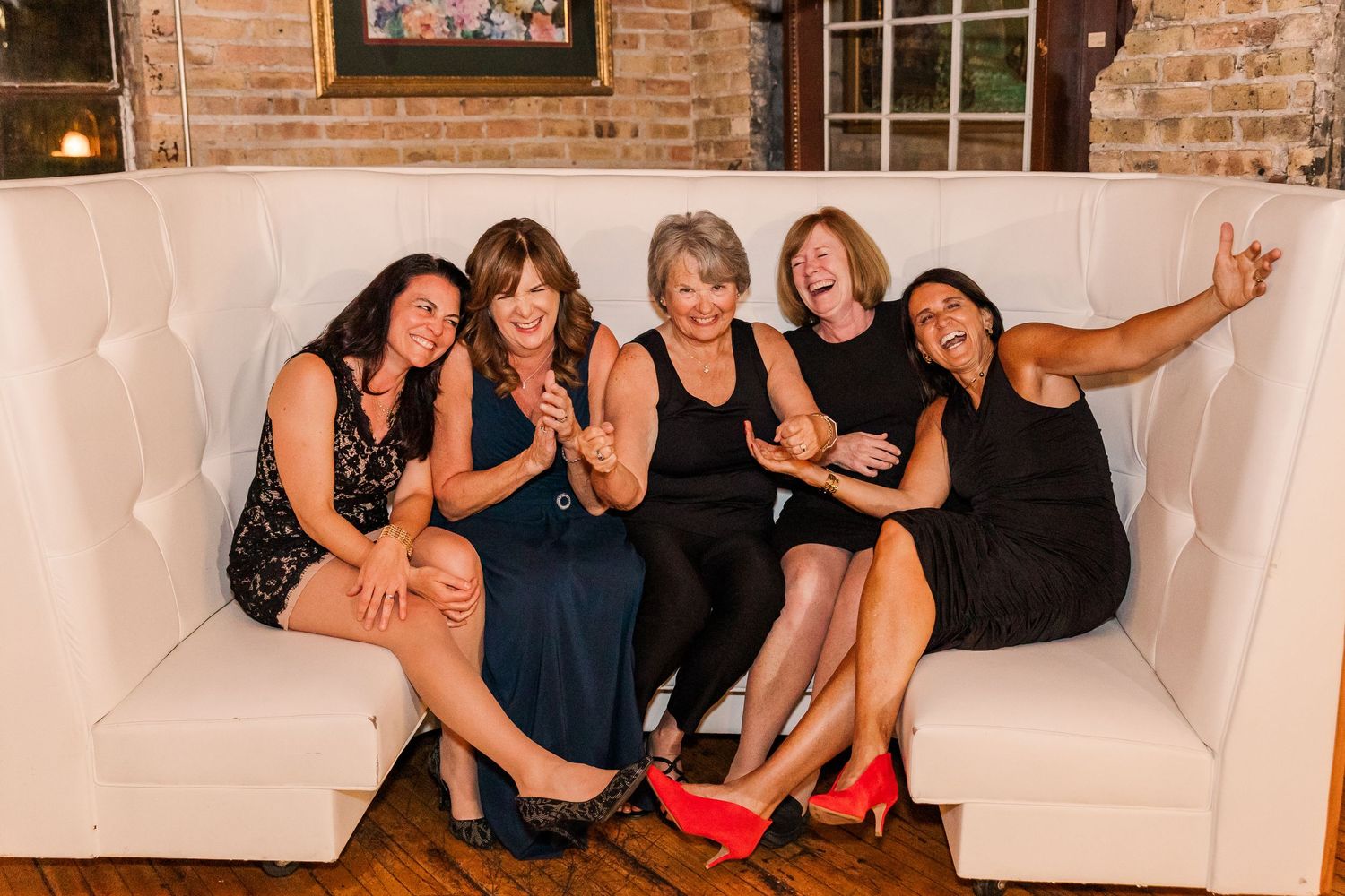 A group of wedding guests in black dresses pose together on a white couch at a rustic brick venue.