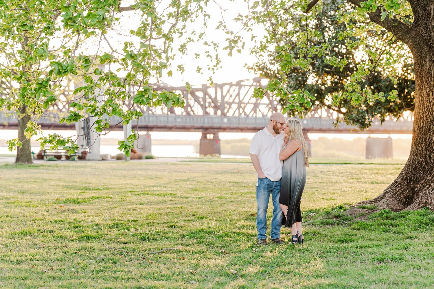 A couple walks together under blooming trees near a bridge at sunset.