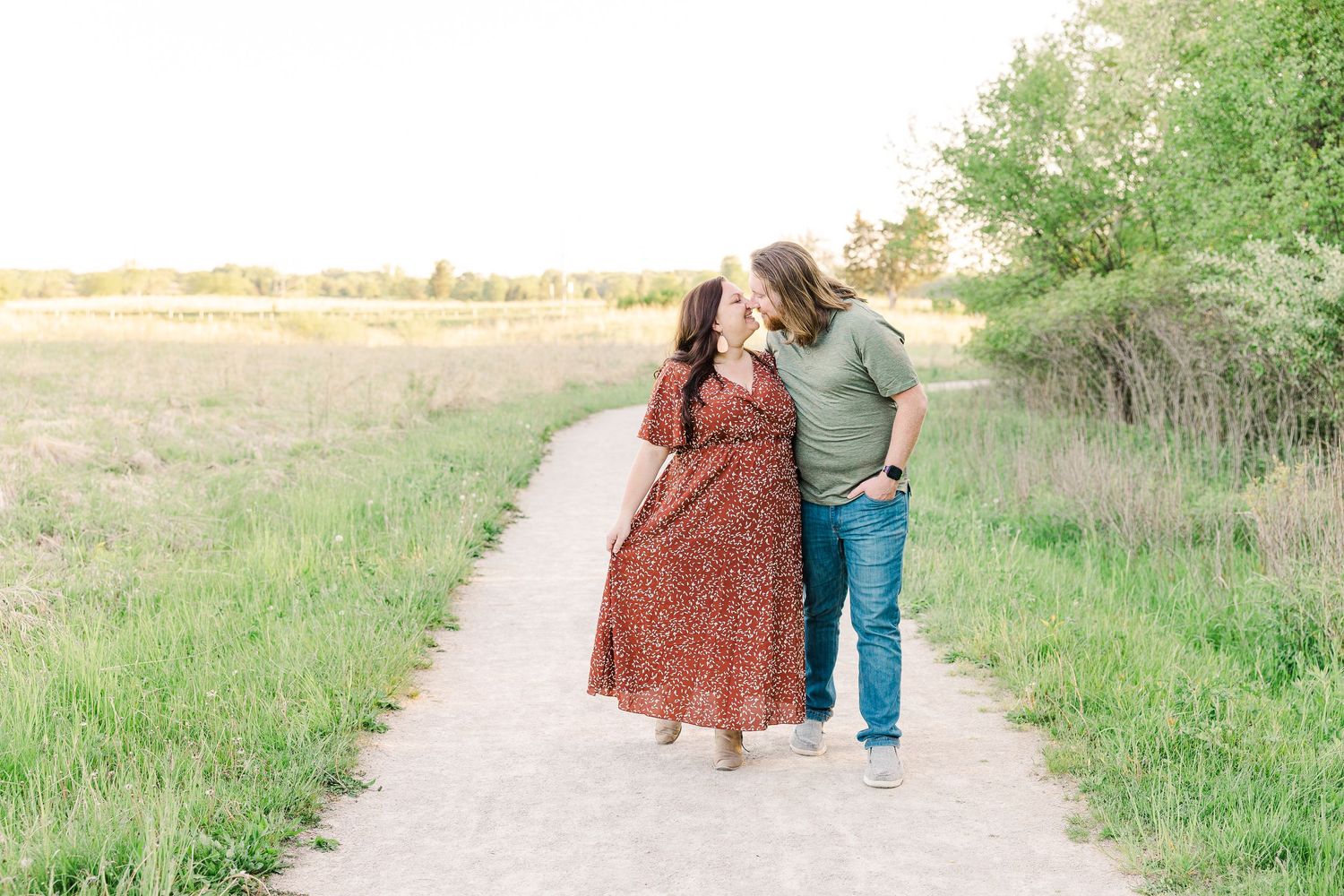 Two people sharing a romantic moment while walking along a sunlit dirt path through green fields.