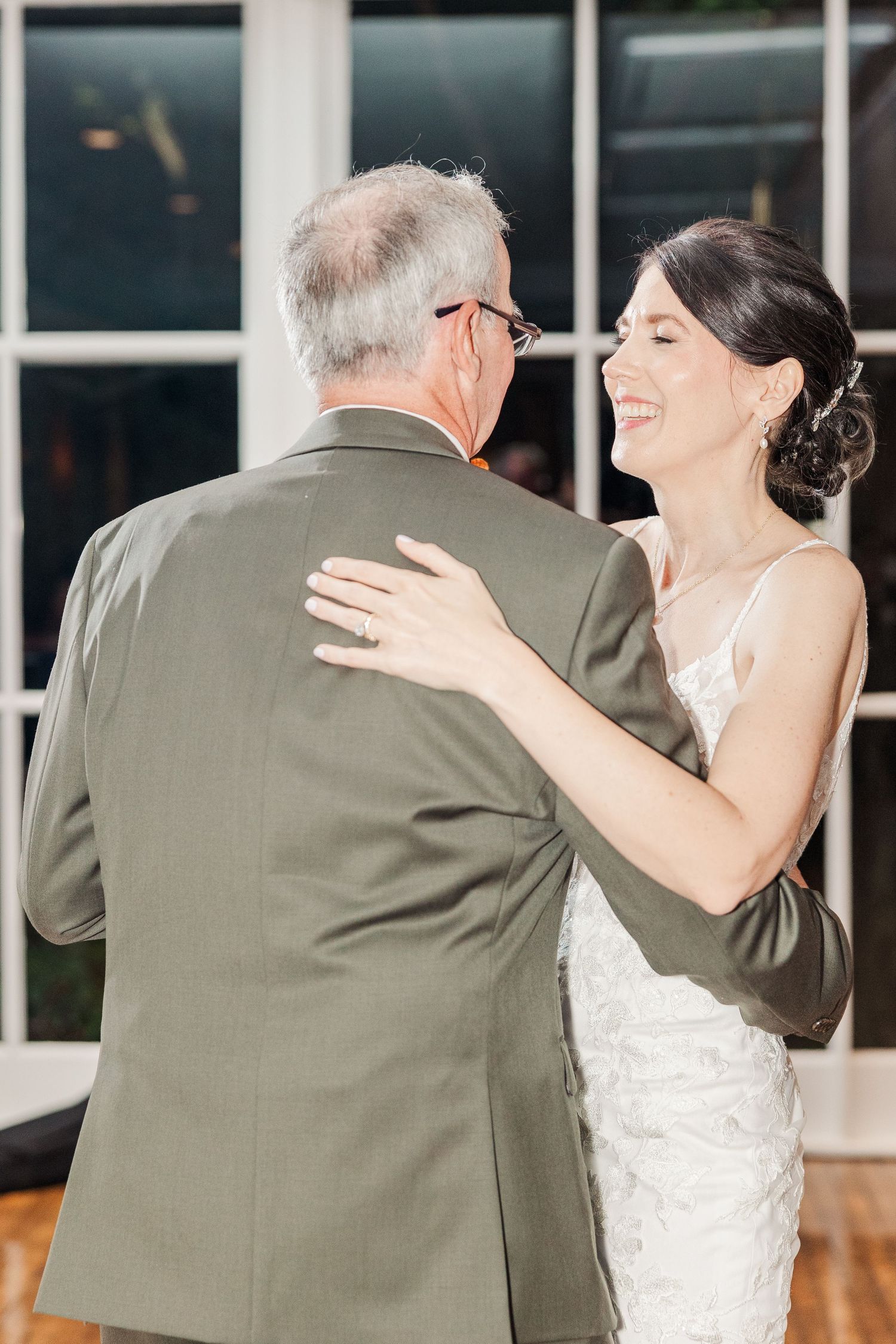 A heartfelt dance between a bride in a white lace gown and an older gentleman in a grey suit at a wedding reception.