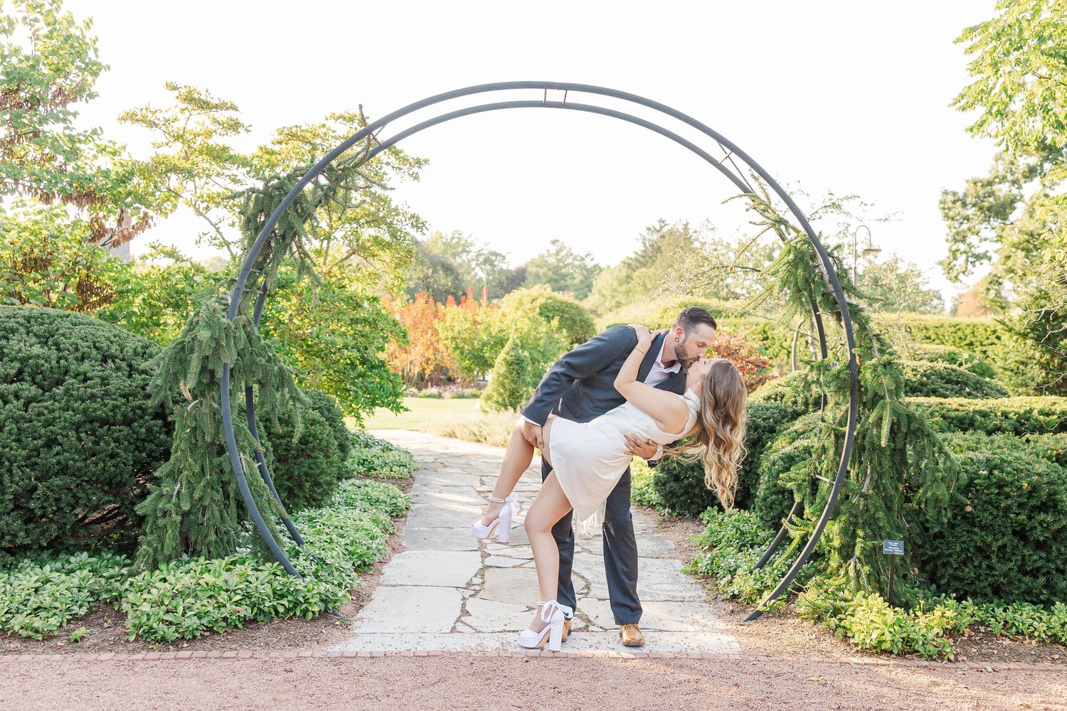 A couple walks along a garden path surrounded by lush greenery on a sunny afternoon.