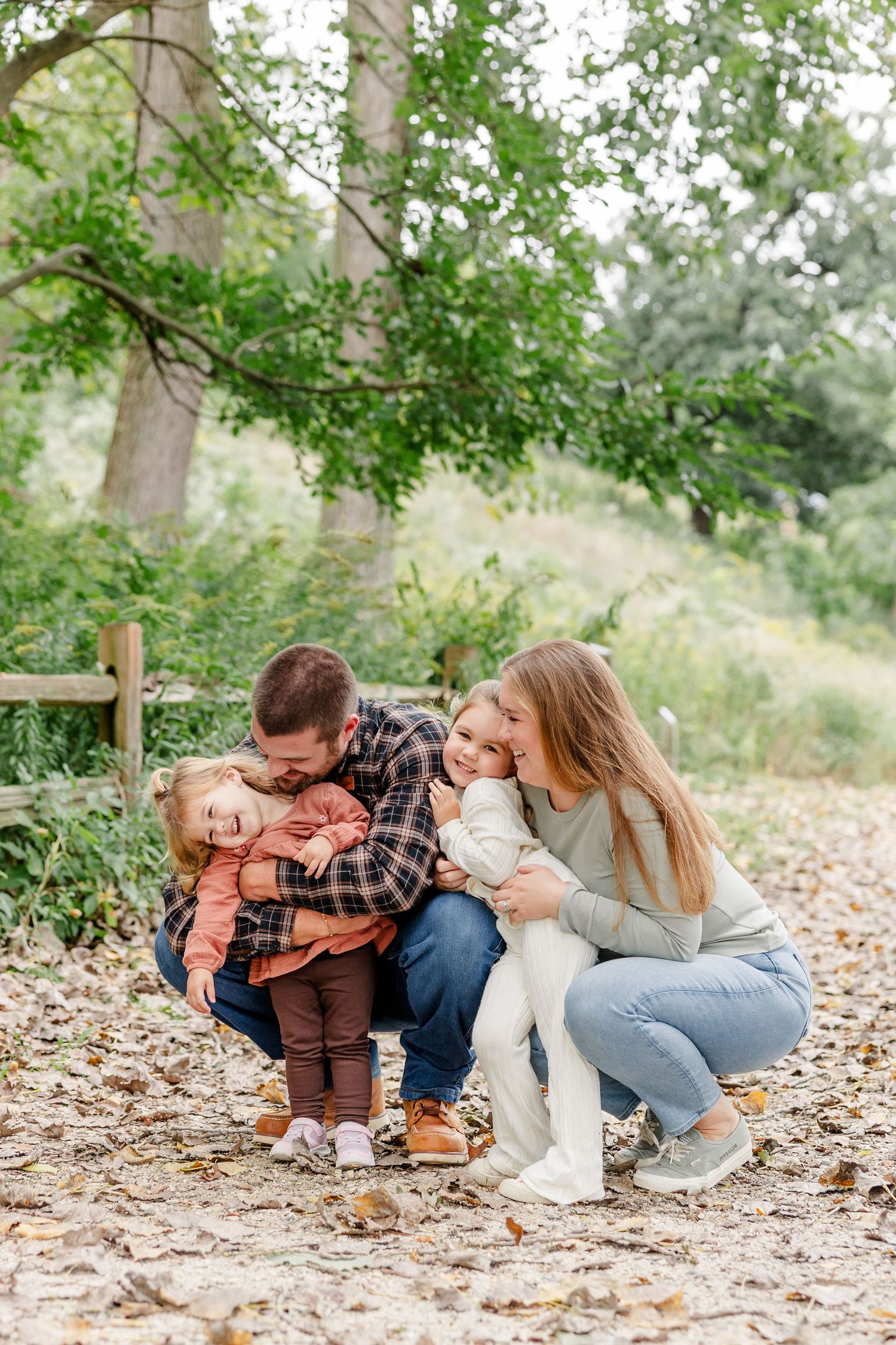 A happy family of four crouches together on a leaf-covered path in an autumn park, parents kissing their two young children.
