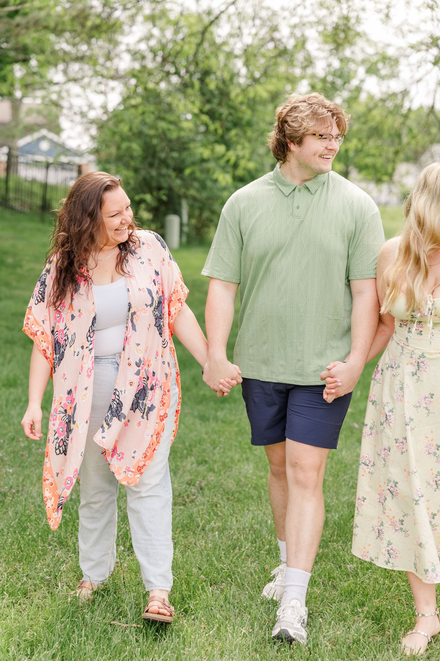 A woman and teenage boy hold hands walking on green grass, wearing casual spring outfits outdoors.